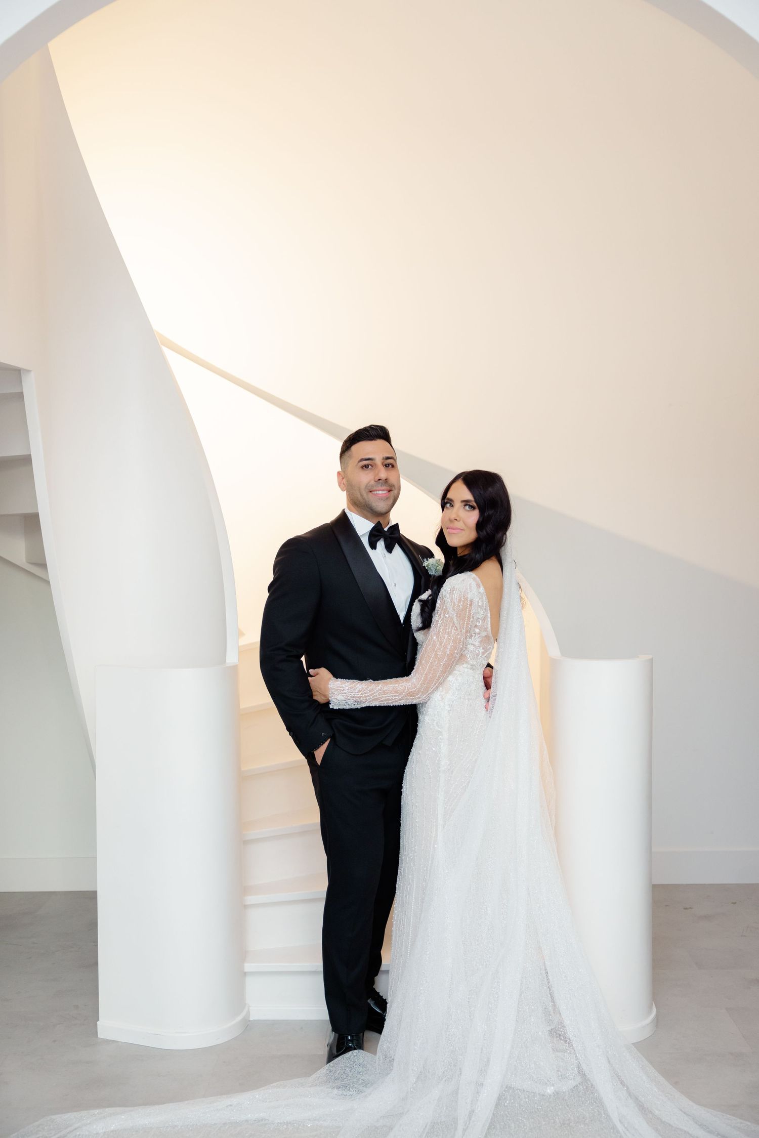 Stylish bride and groom embracing by curved white staircase in contemporary setting.
