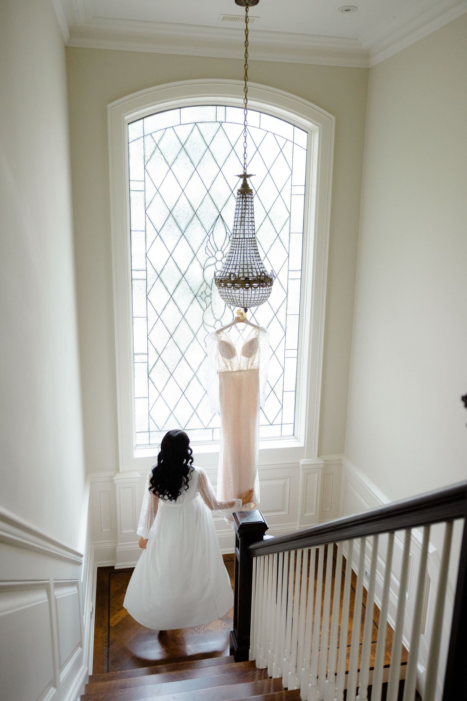 Elegant stairwell scene with wedding gown displayed beneath crystal chandelier by arched window.