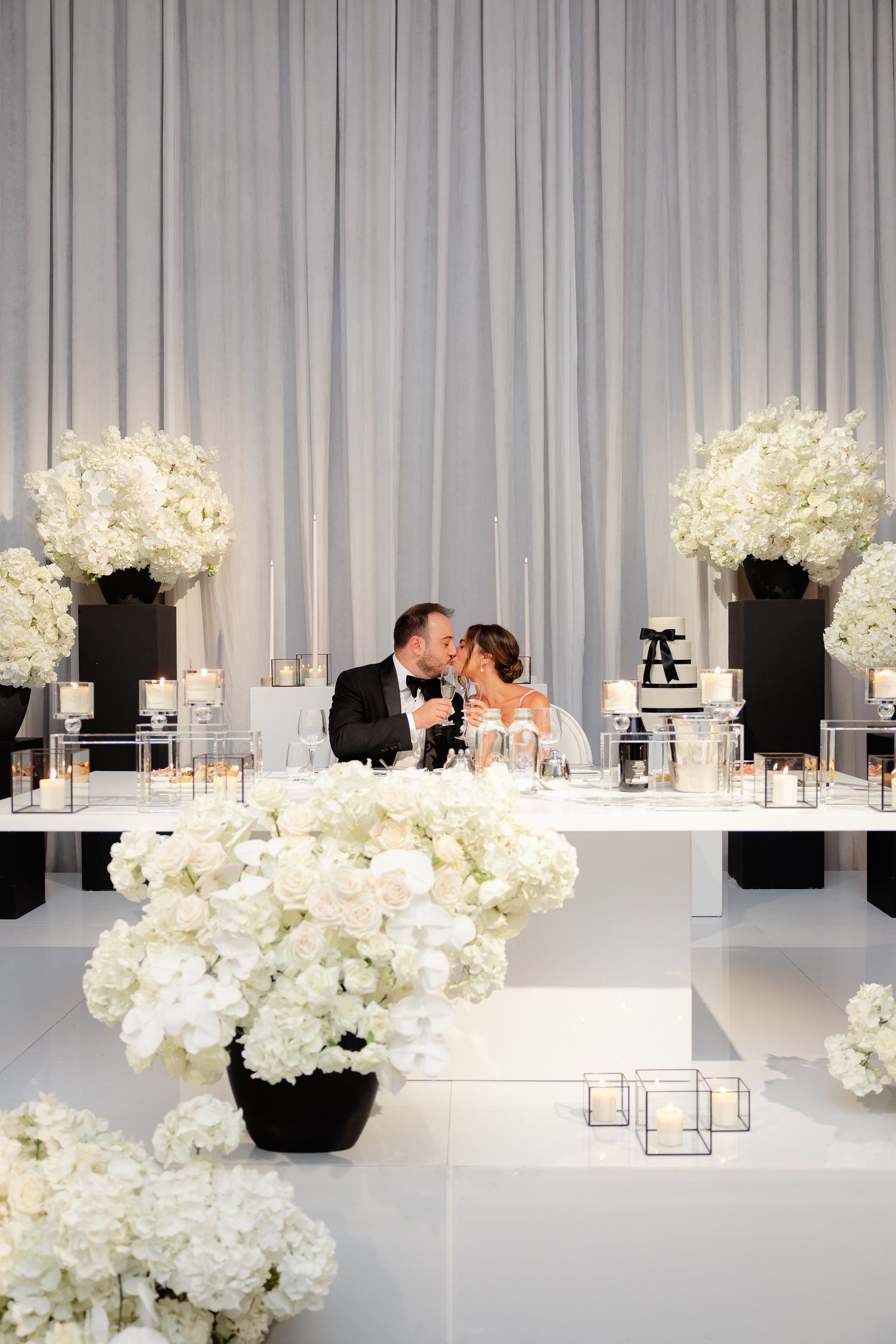 Elegant white wedding reception decor with hydrangeas in black vases and candles against a gray curtain backdrop.