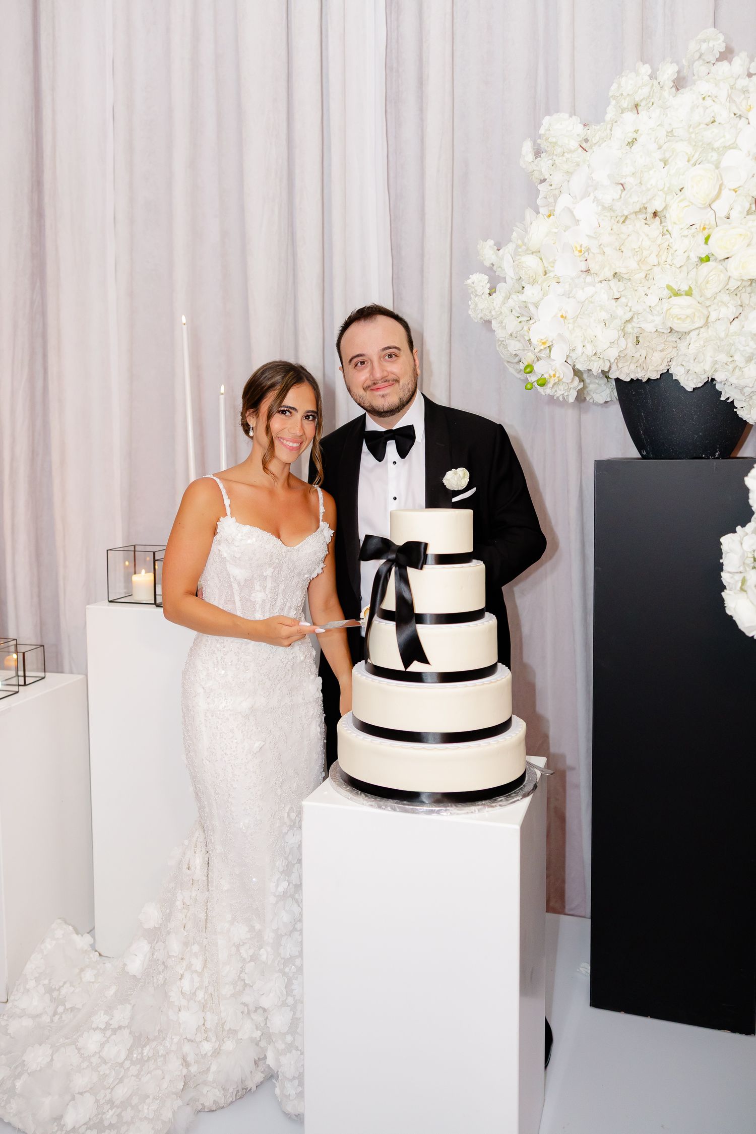Elegant black and white wedding cake displayed on white pedestal with white floral arrangements and drapery in background.