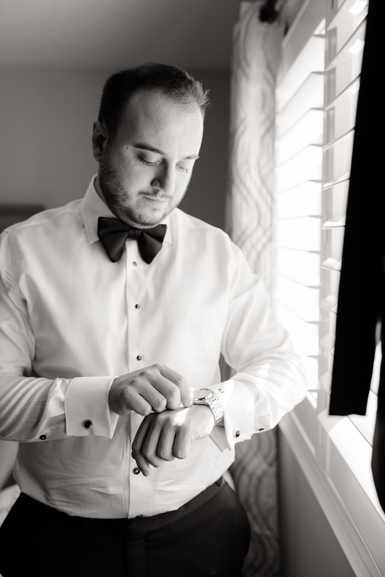 A person in a white dress shirt and bow tie checks their watch while standing near a window in black and white.