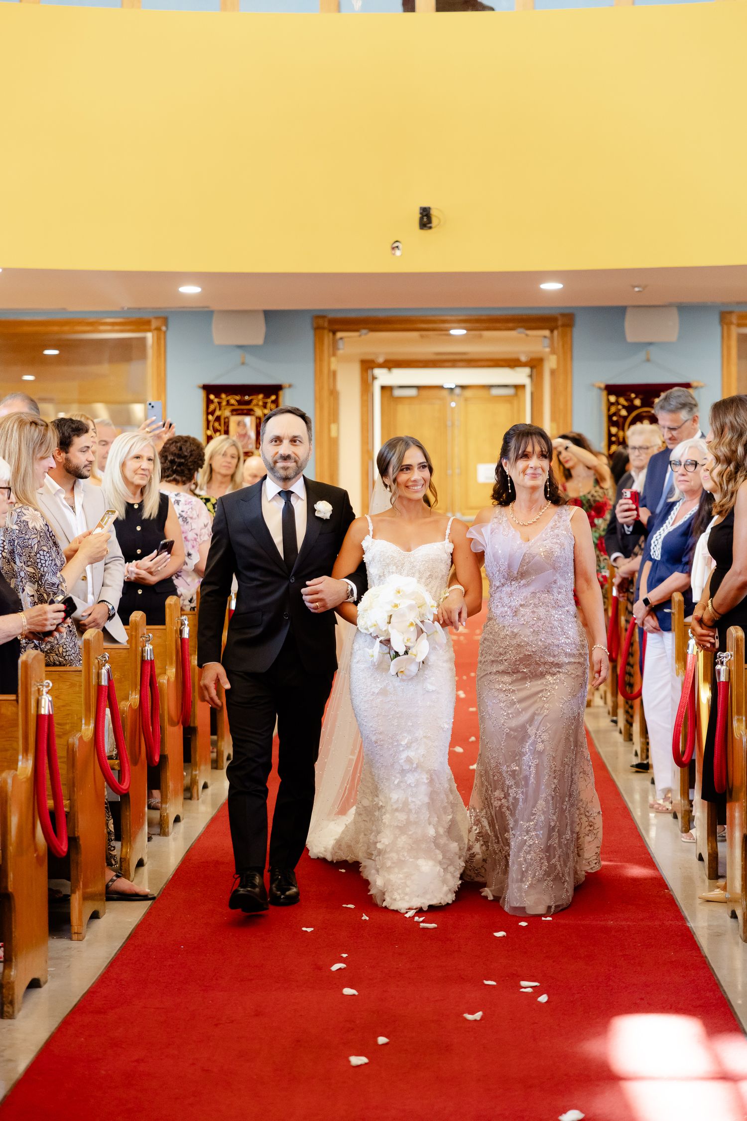 Wedding party walks down red-carpeted church aisle while guests look on during ceremony.