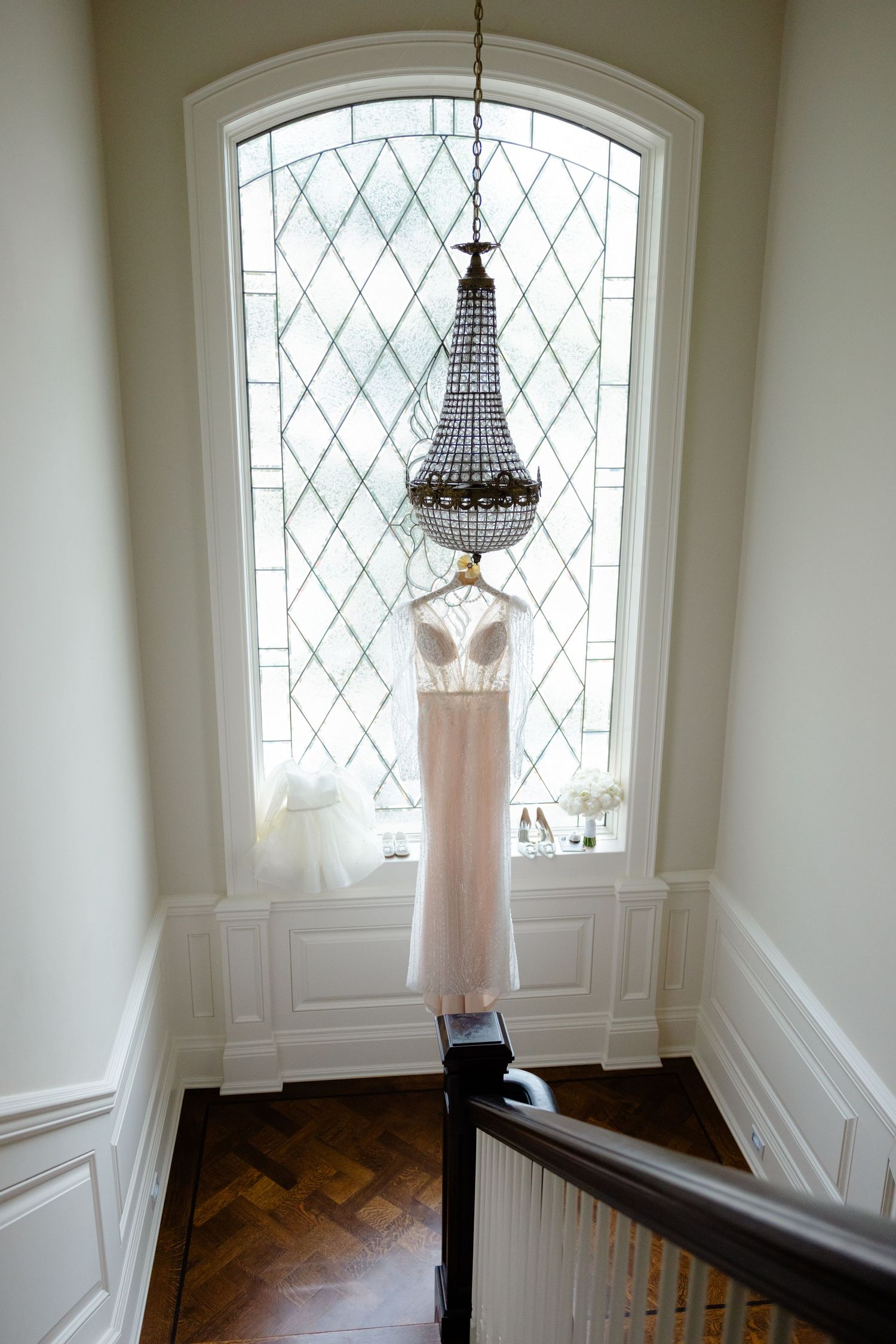 Blush wedding gown hanging beneath crystal chandelier in elegant window alcove.