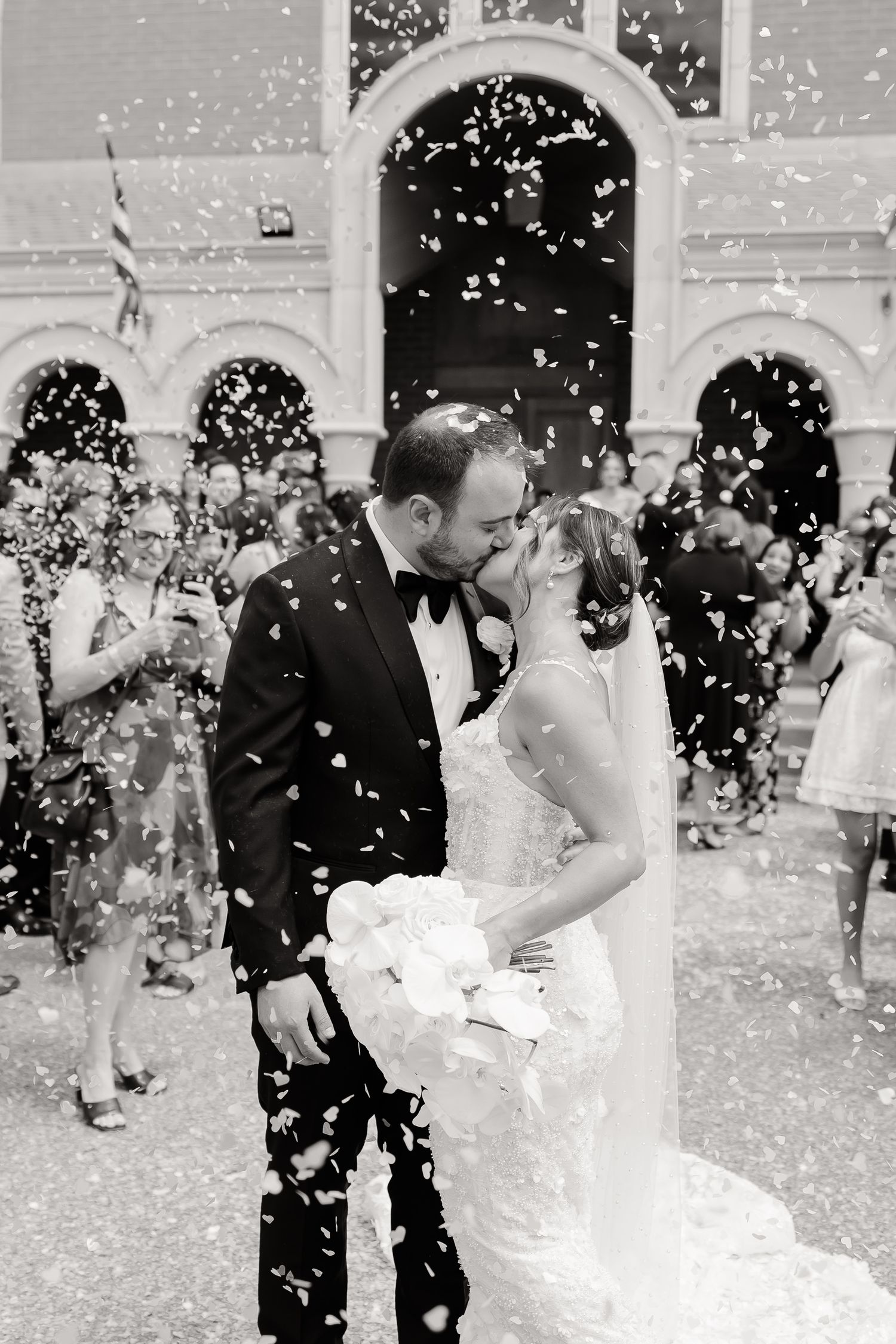 Wedding couple shares a kiss outside a church while guests throw white confetti in a joyful black and white moment.