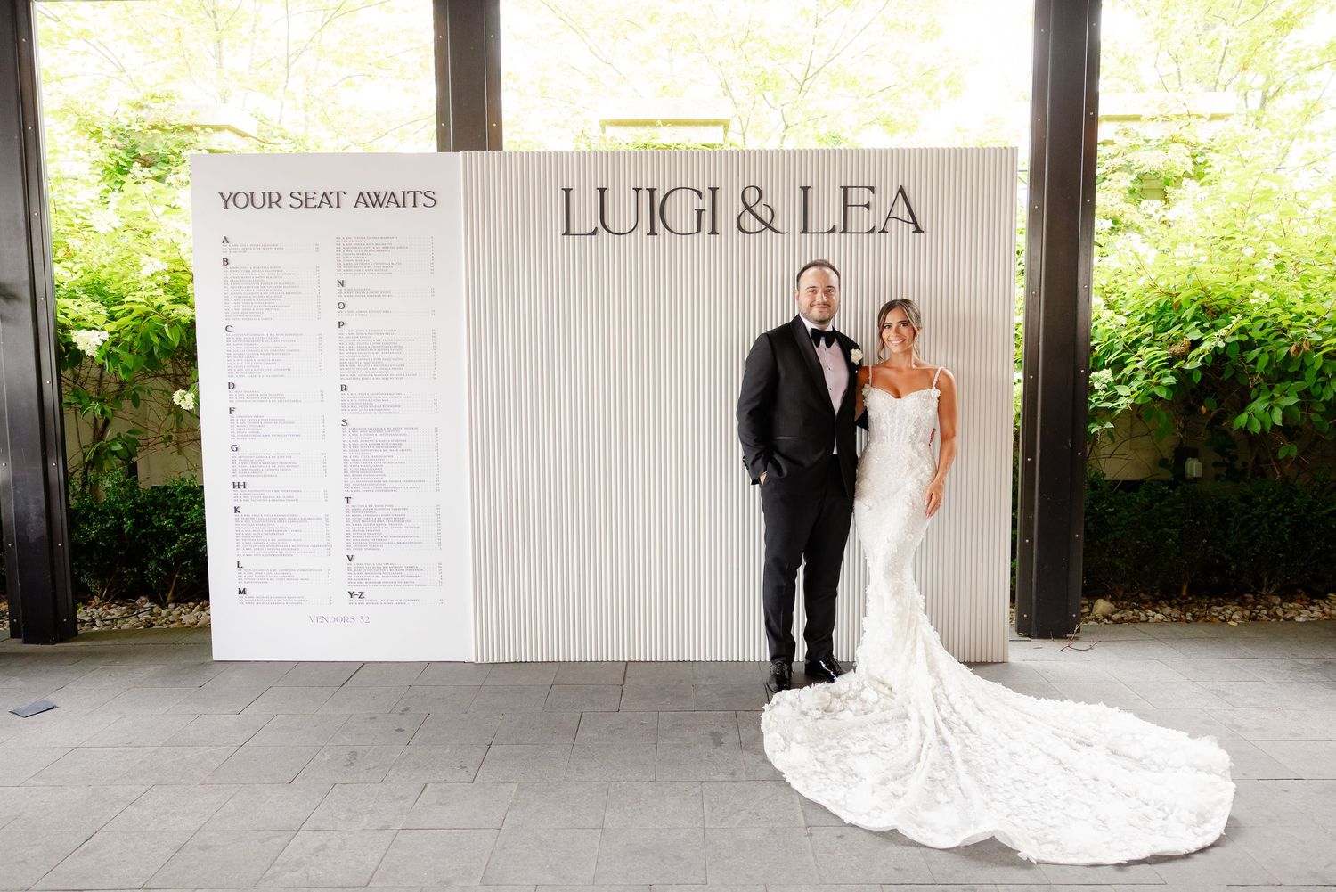 A couple poses in formal attire in front of a white backdrop with elegant seating chart at a wedding reception.