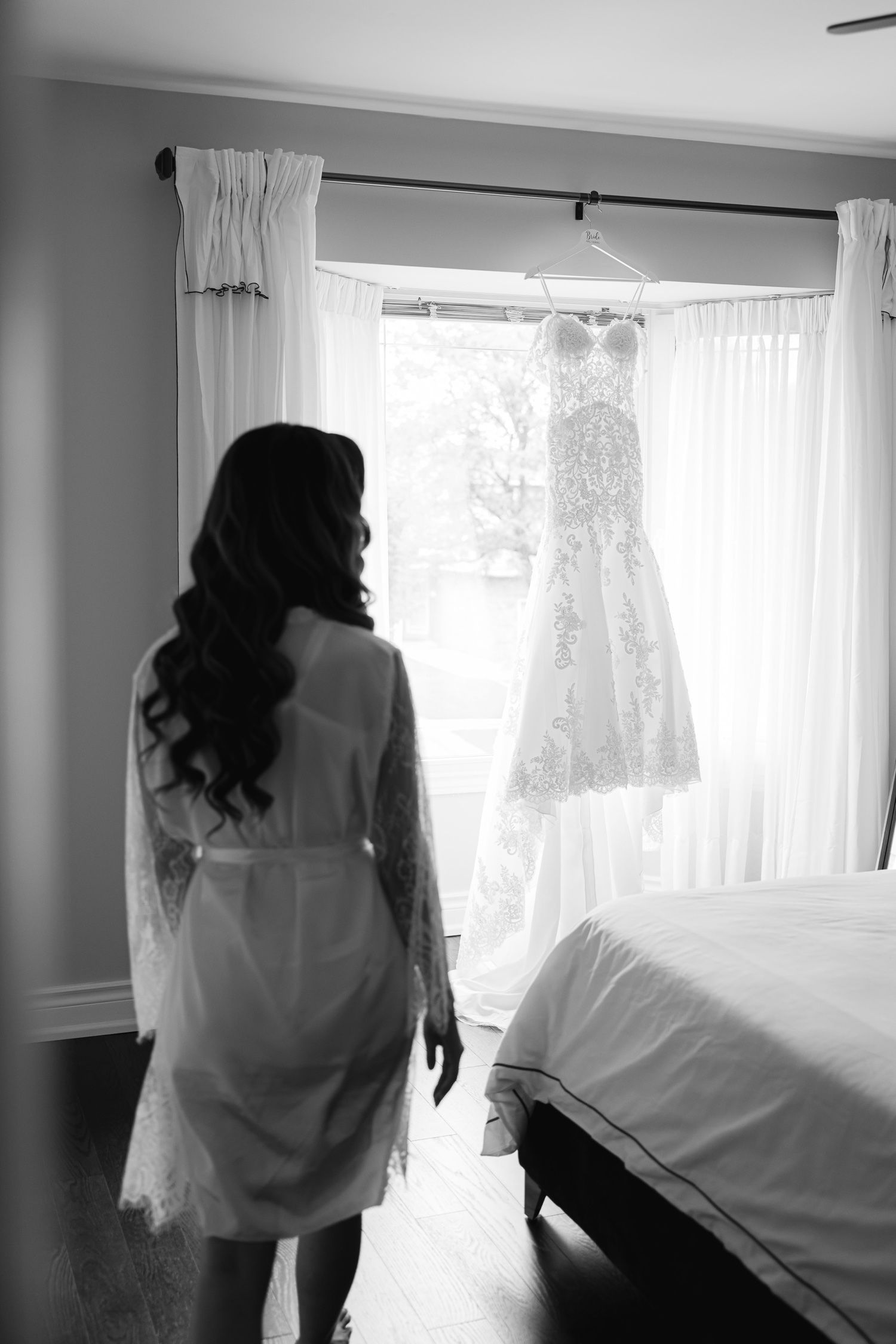 Silhouette of person in white robe looking at wedding dress hanging by window.