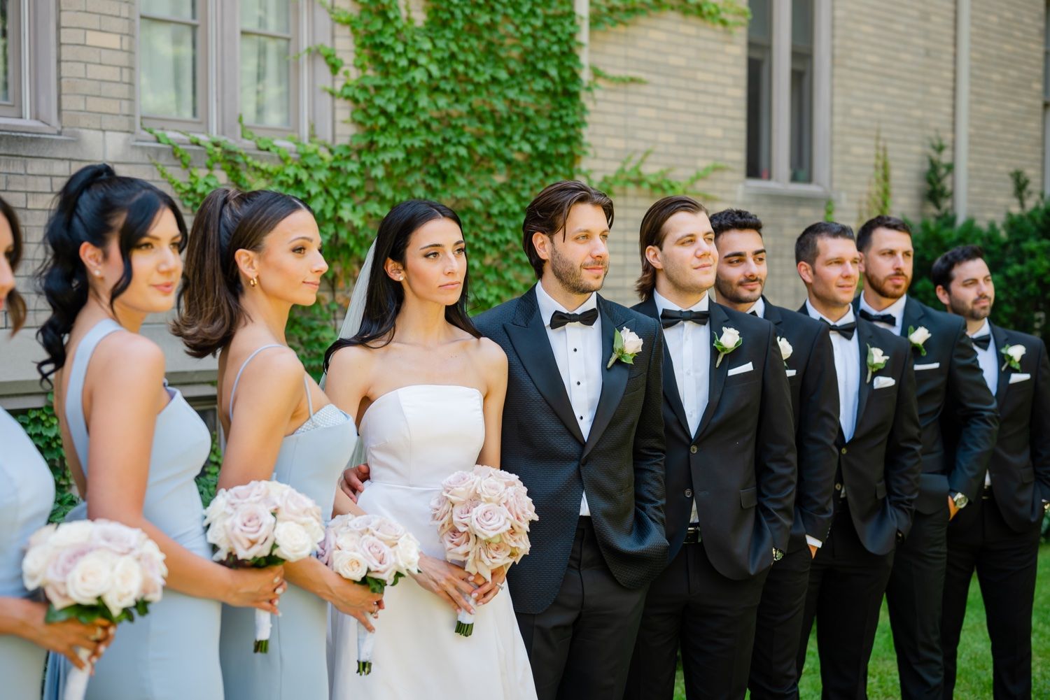 Wedding party lines up outdoors wearing blue bridesmaid dresses and black tuxedos against an ivy-covered wall.