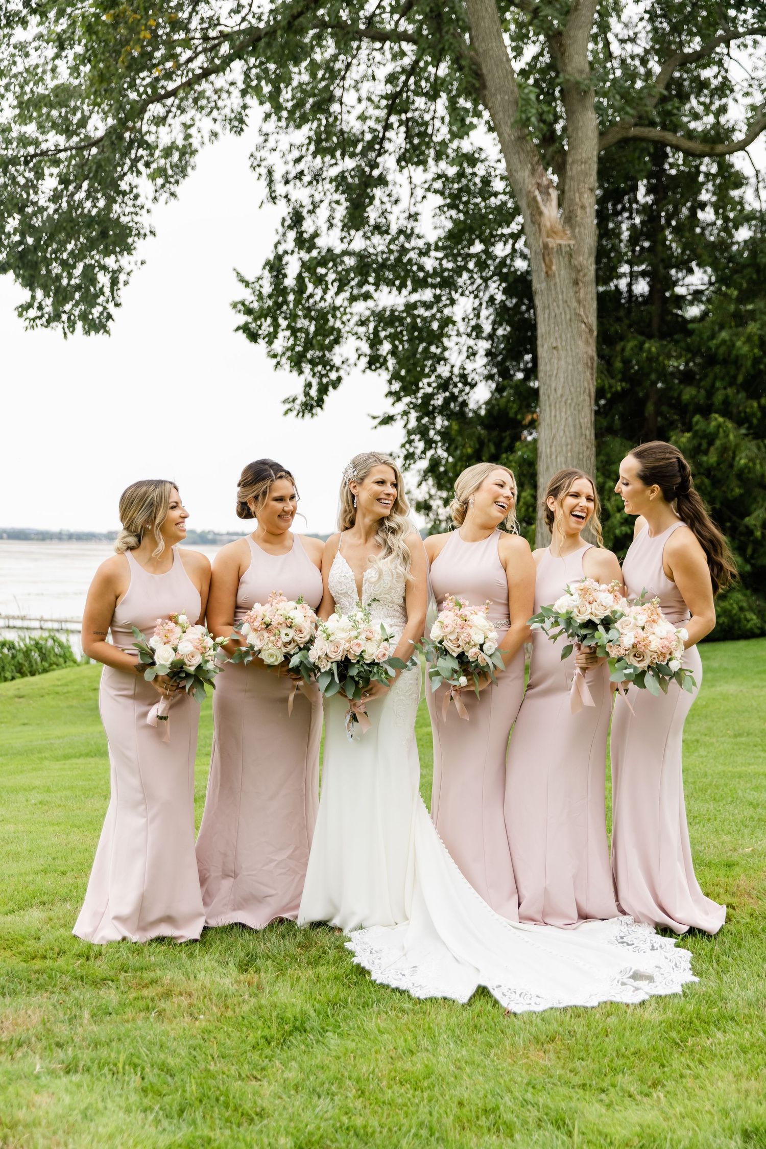 Wedding party in blush pink bridesmaid dresses poses with bride in flowing white gown under a large tree by the water.
