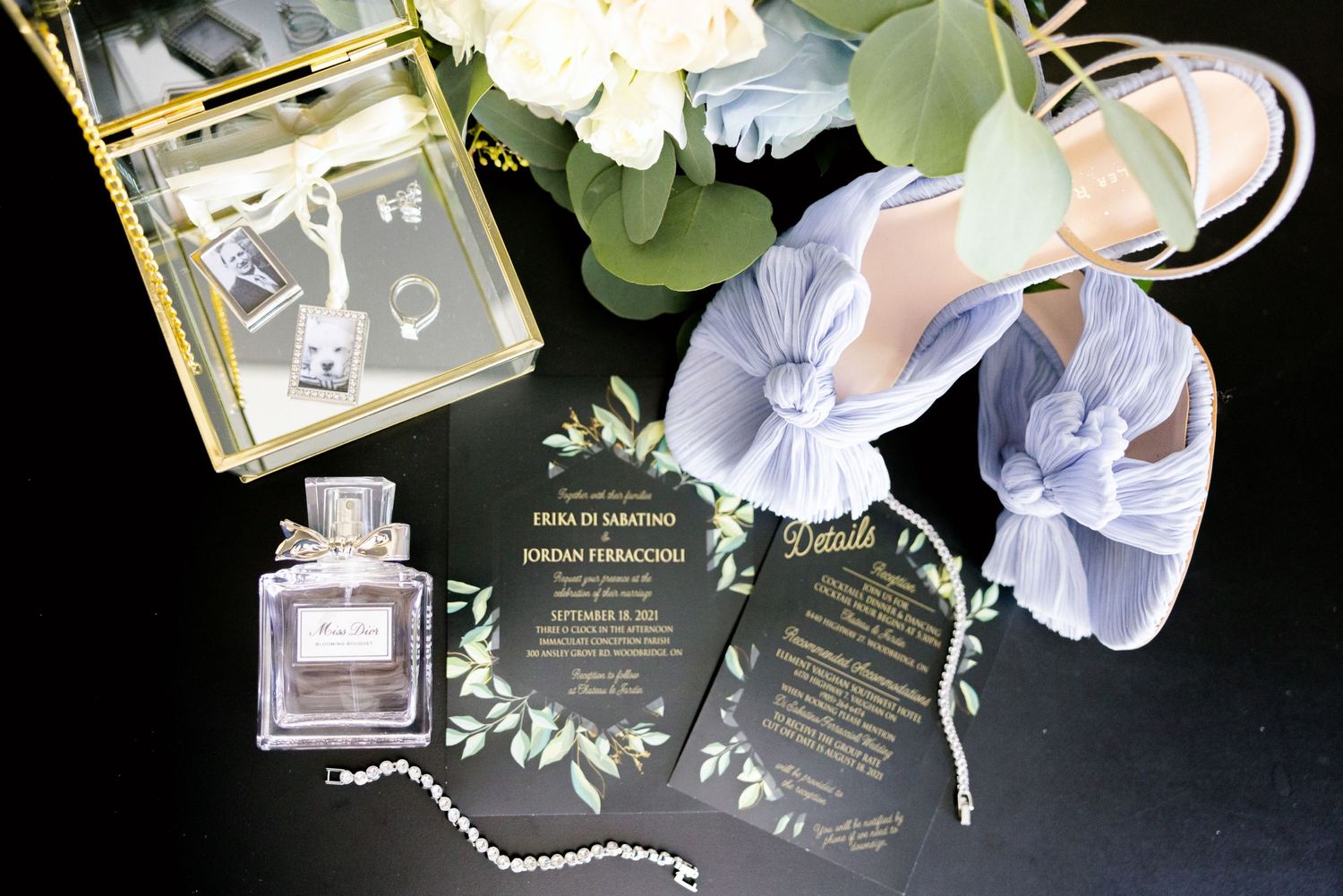 Wedding details flatlay with perfume bottle, invitations, and light blue shoes against dark background.