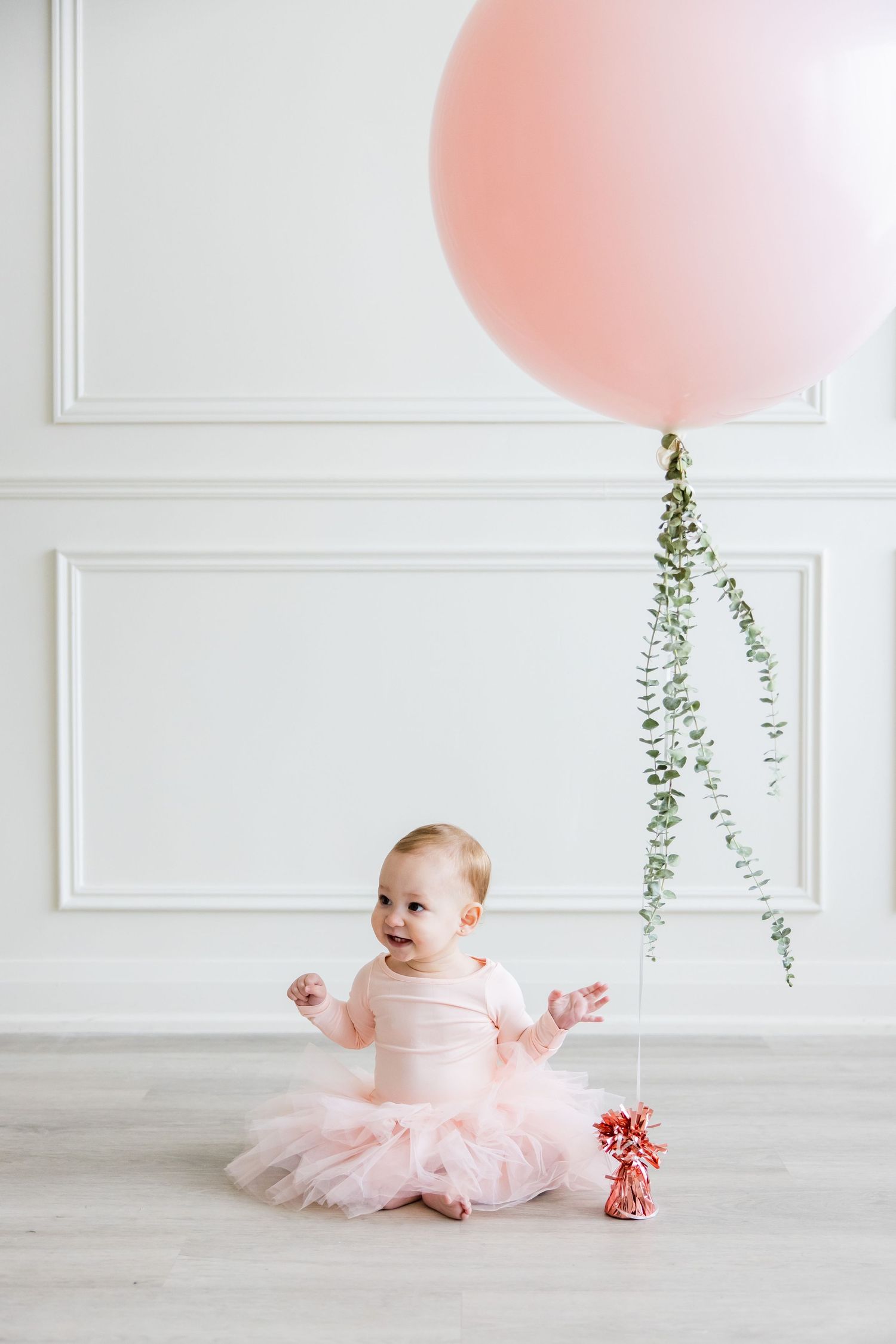 Simple first birthday setup with pink balloon, white wall panels, and hanging greenery.