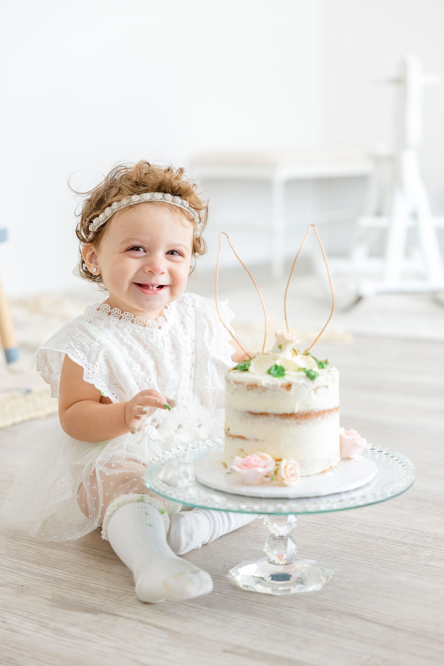 A child in white lace smiles brightly next to an elegant cake topped with wire bunny ears.