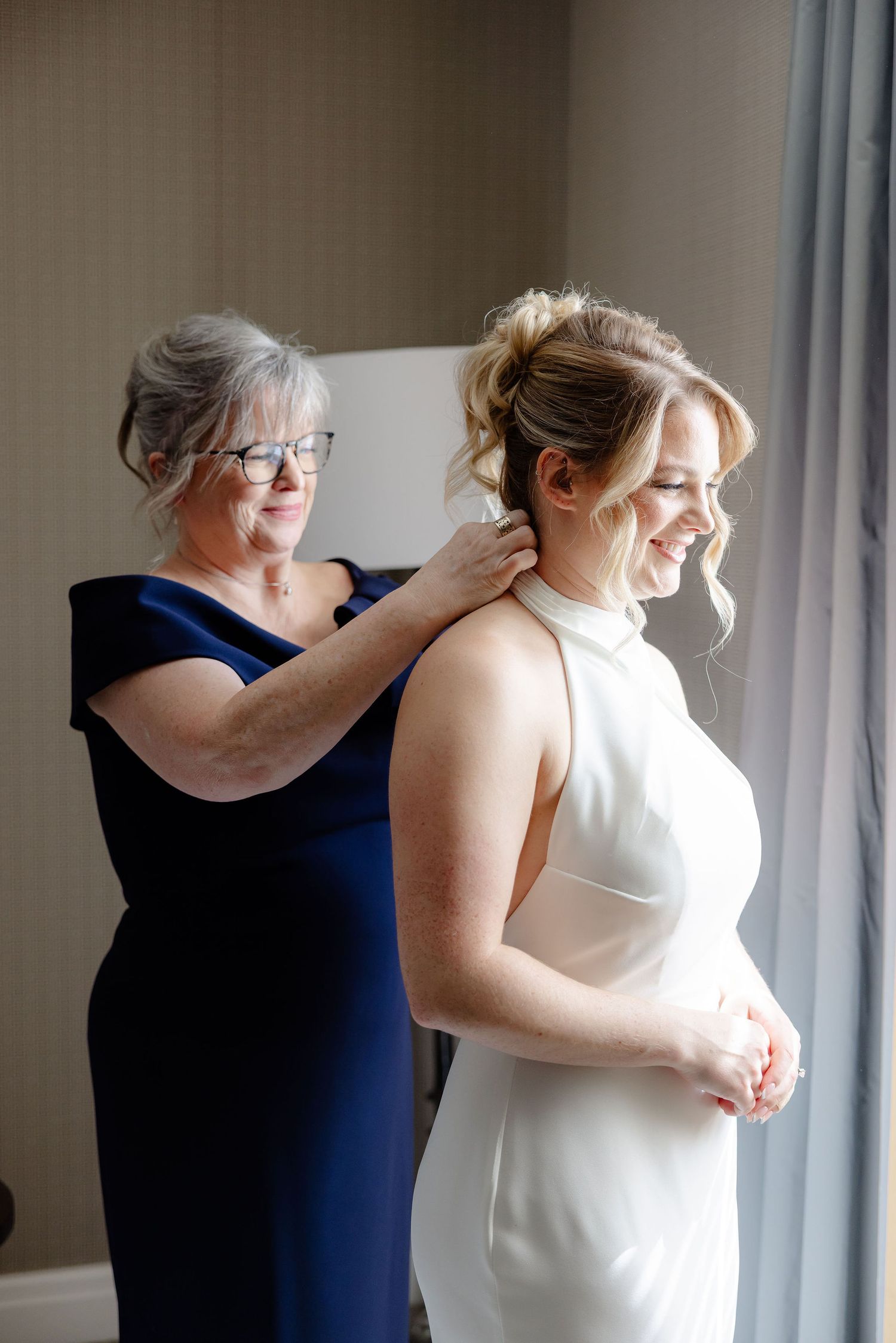 A bride in a white dress gets help with final adjustments from someone wearing navy blue in a bright room.