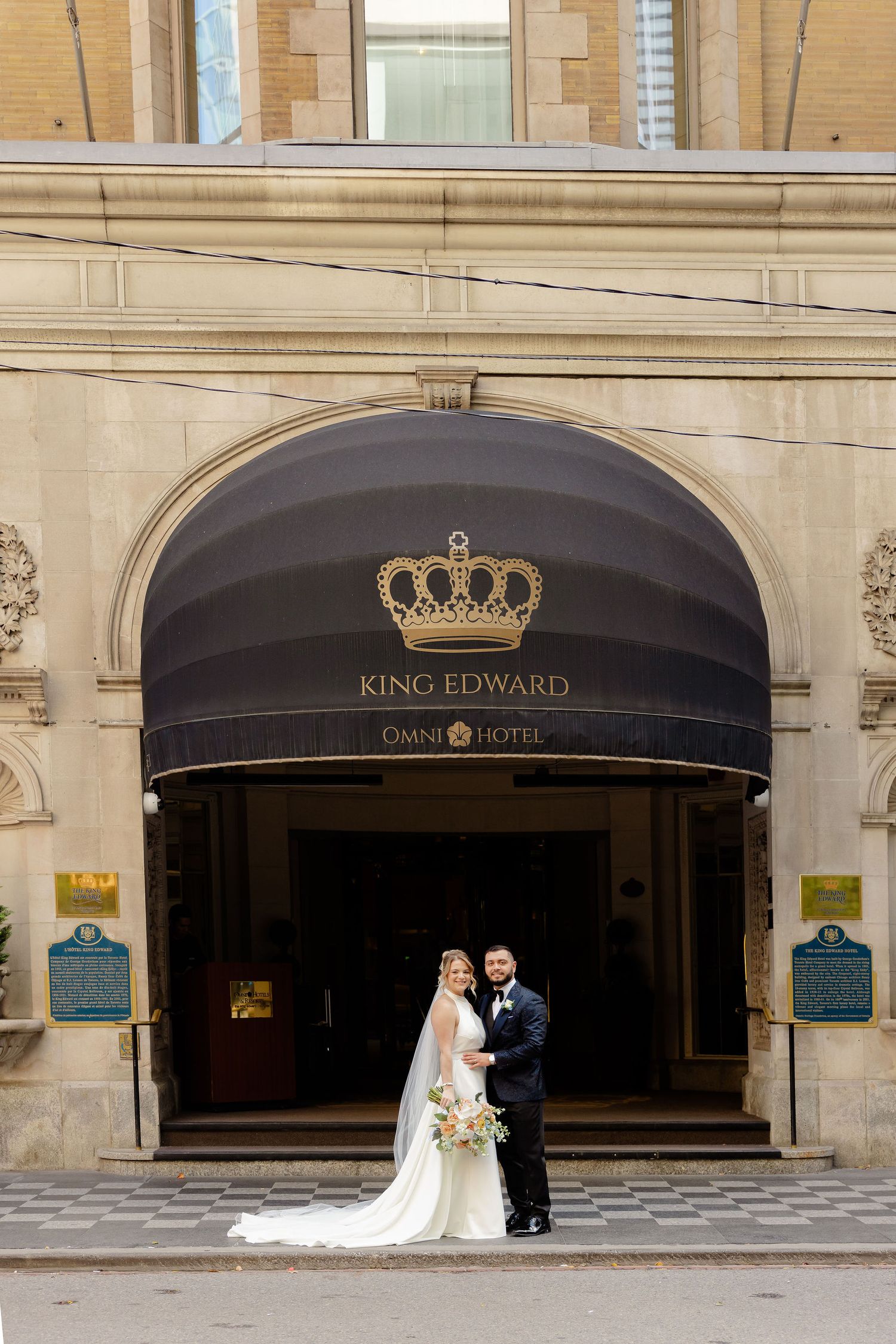 A wedding couple poses outside the elegant King Edward Hotel entrance with black awnings and stone facade.