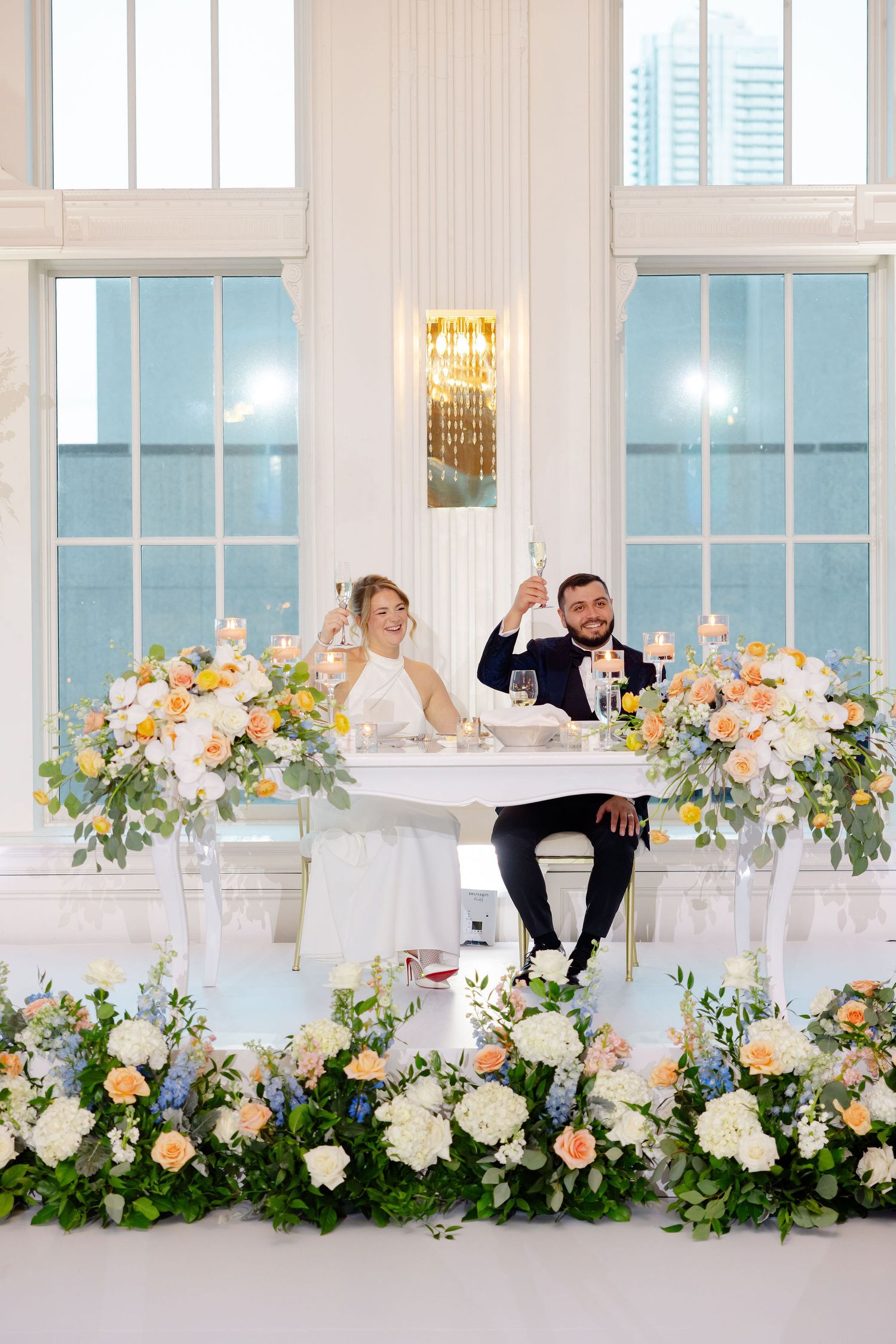Wedding couple celebrates at decorated sweetheart table with champagne toast against bright windows.