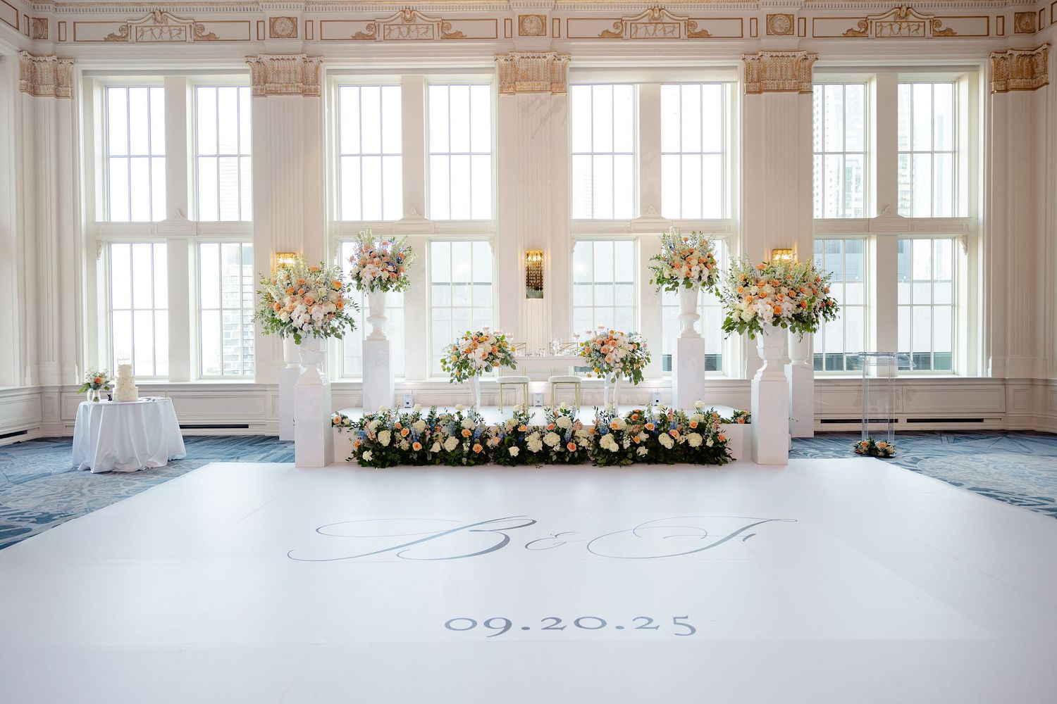 Elegant white ballroom with ornate moldings and large windows features romantic floral arrangements for a wedding celebration.