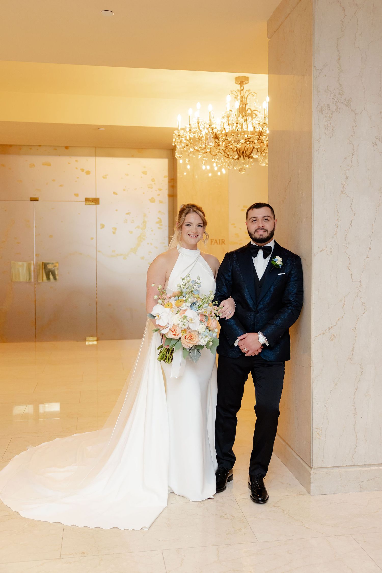 Wedding couple poses playfully in formal attire against marble walls and chandeliers.