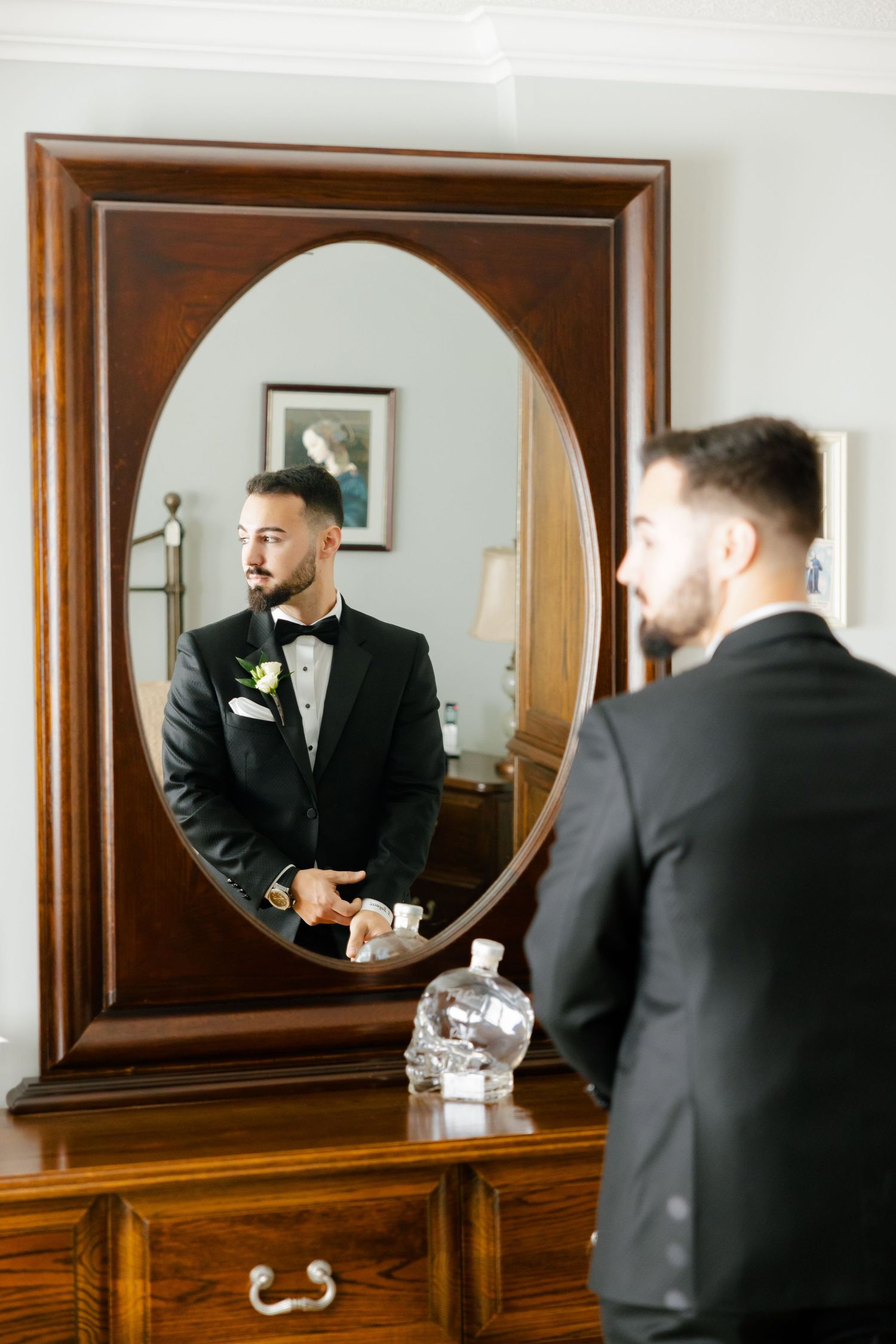 A reflection in an ornate mirror shows a groom adjusting his bow tie before the wedding ceremony.