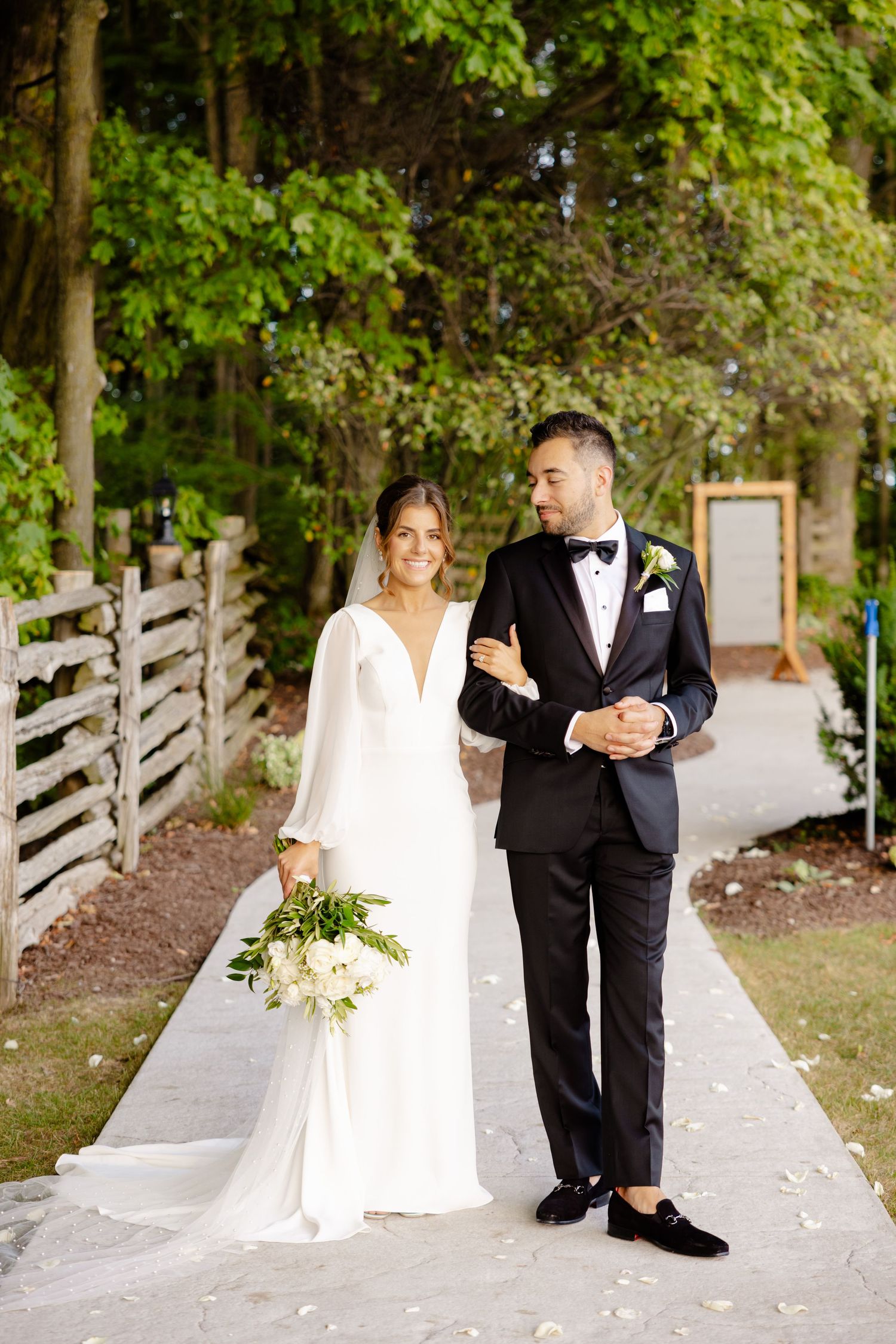 A formal wedding portrait on a garden path with the bride holding a white floral bouquet.