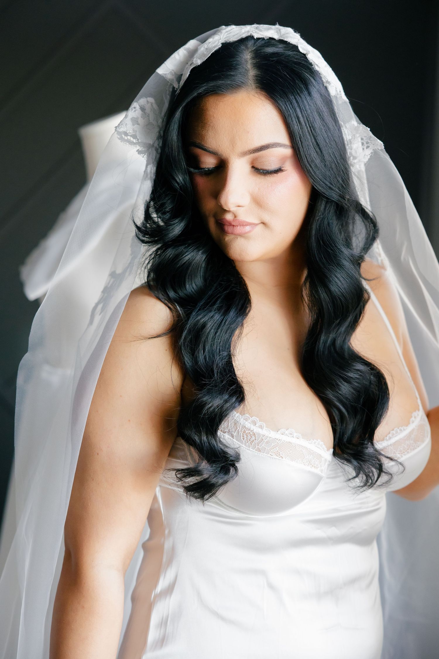 A bride wearing a white silk dress and veil with long dark wavy hair styled for her wedding day.