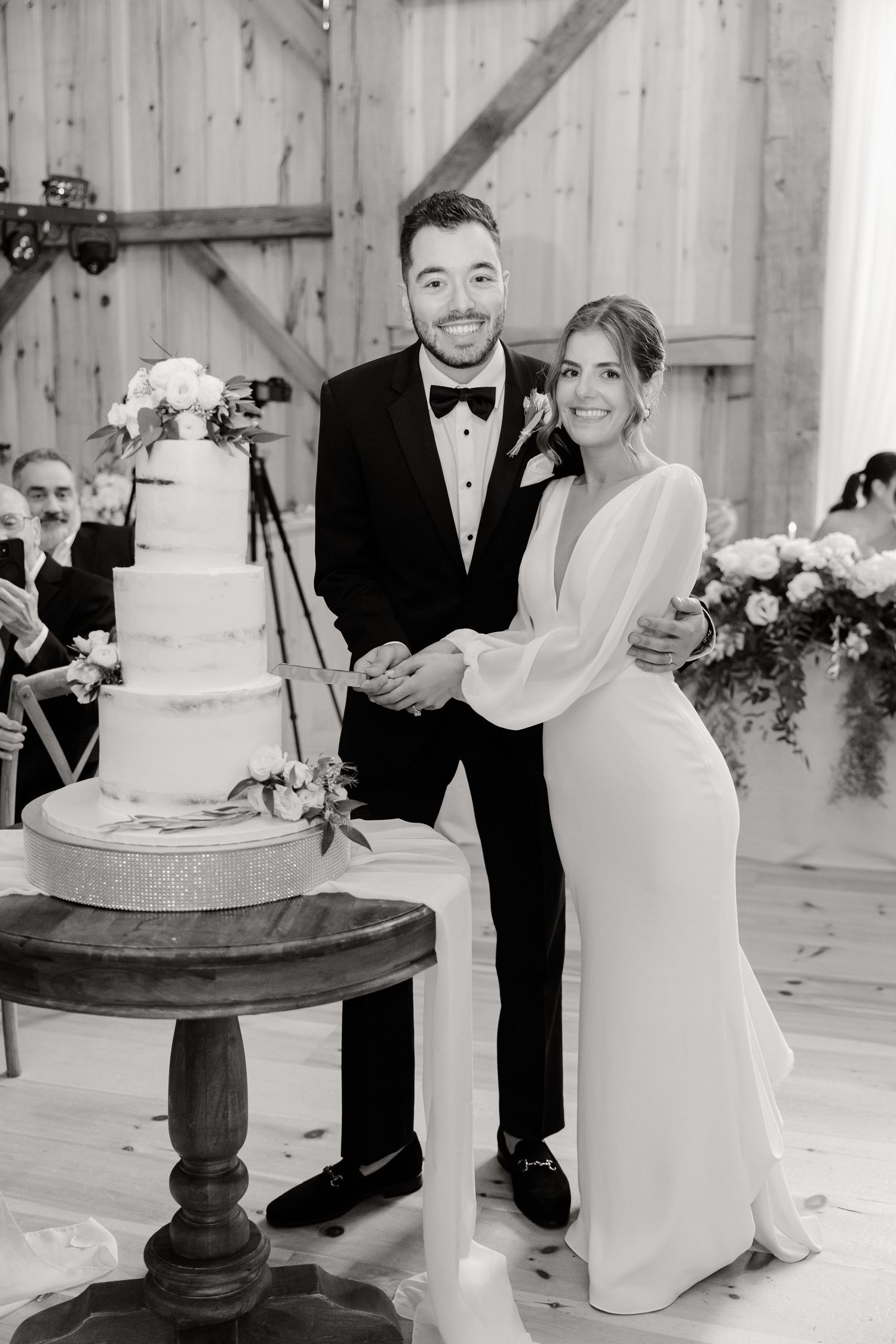 A wedding couple poses beside their elegant white tiered cake in a rustic barn setting.