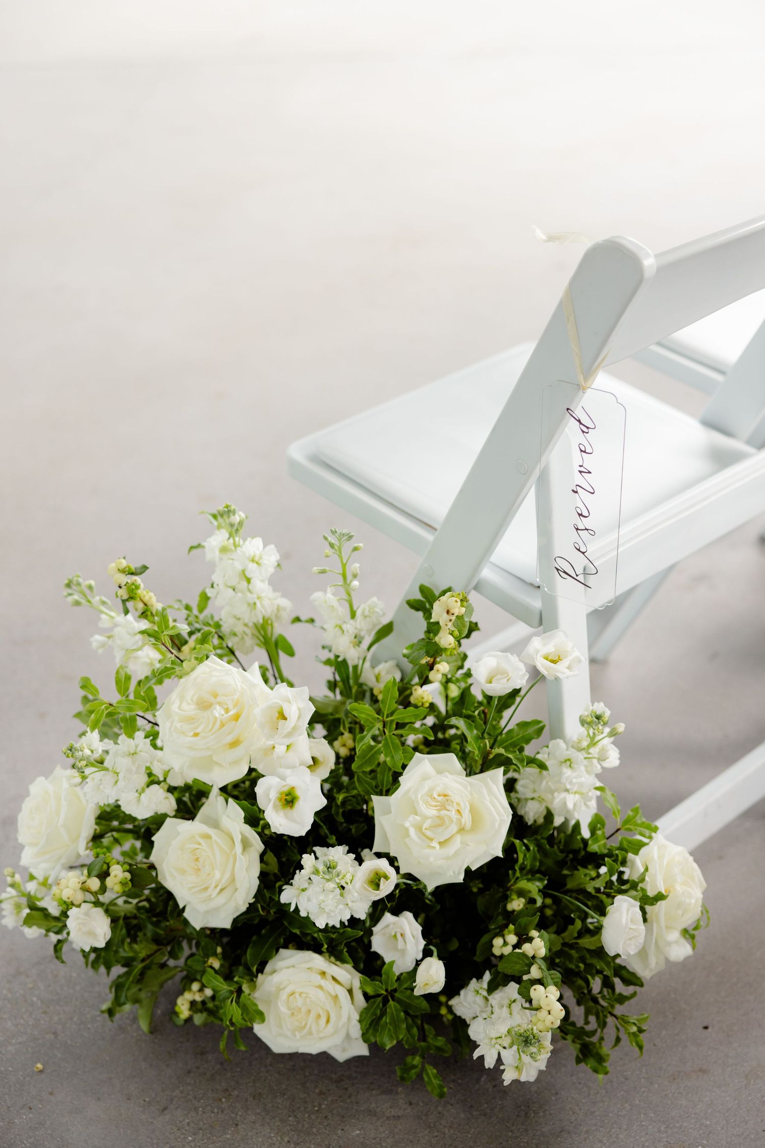 White roses and greenery arranged in a delicate floral display next to a white folding chair.
