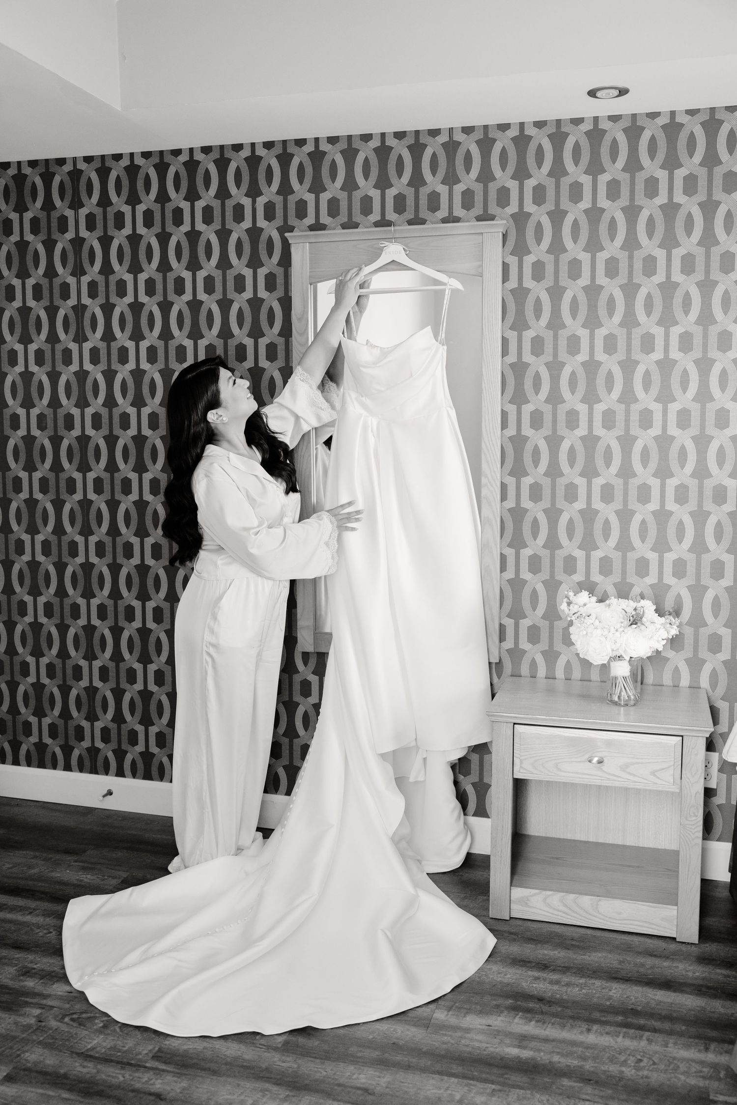 Black and white photo of wedding dress preparation against patterned wall.