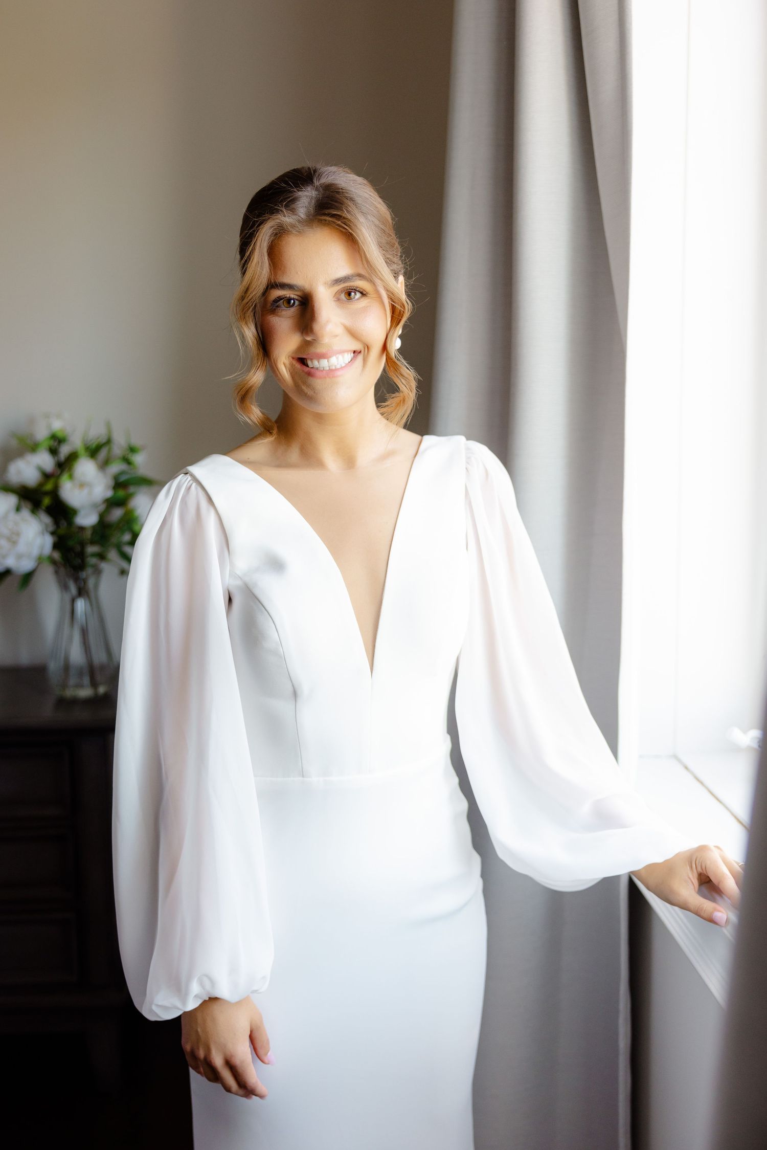 Elegant wedding dress with plunging neckline and billowing sheer sleeves photographed near a window with floral arrangement.