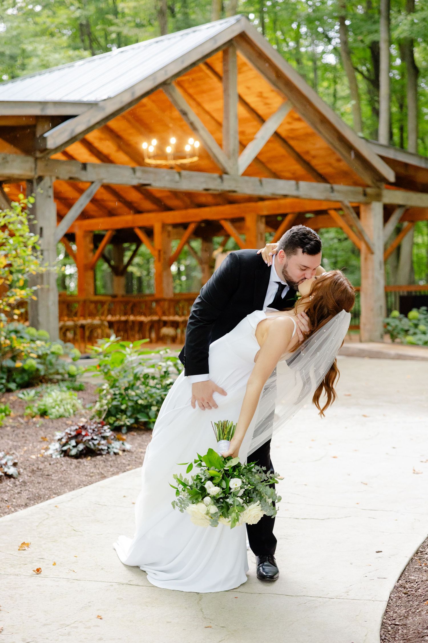 Couple in wedding attire dips romantically beneath wooden pavilion with string lights in wooded garden.