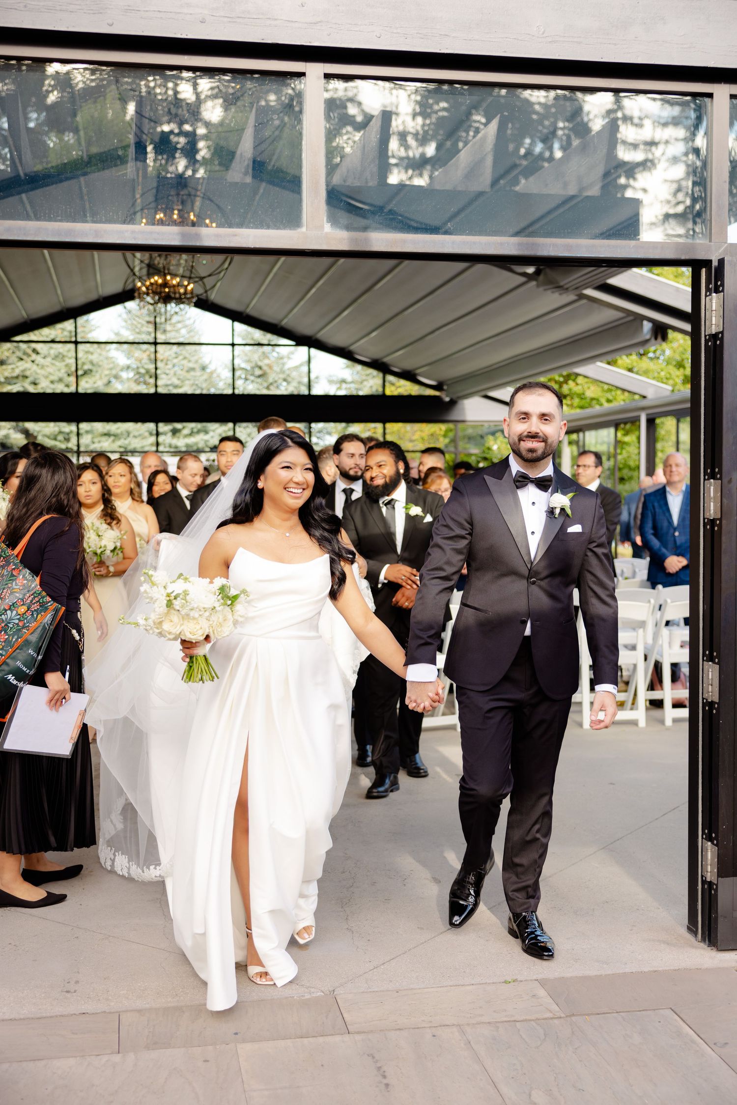 Happy newlyweds exit their ceremony while guests cheer in a modern glass-walled venue.