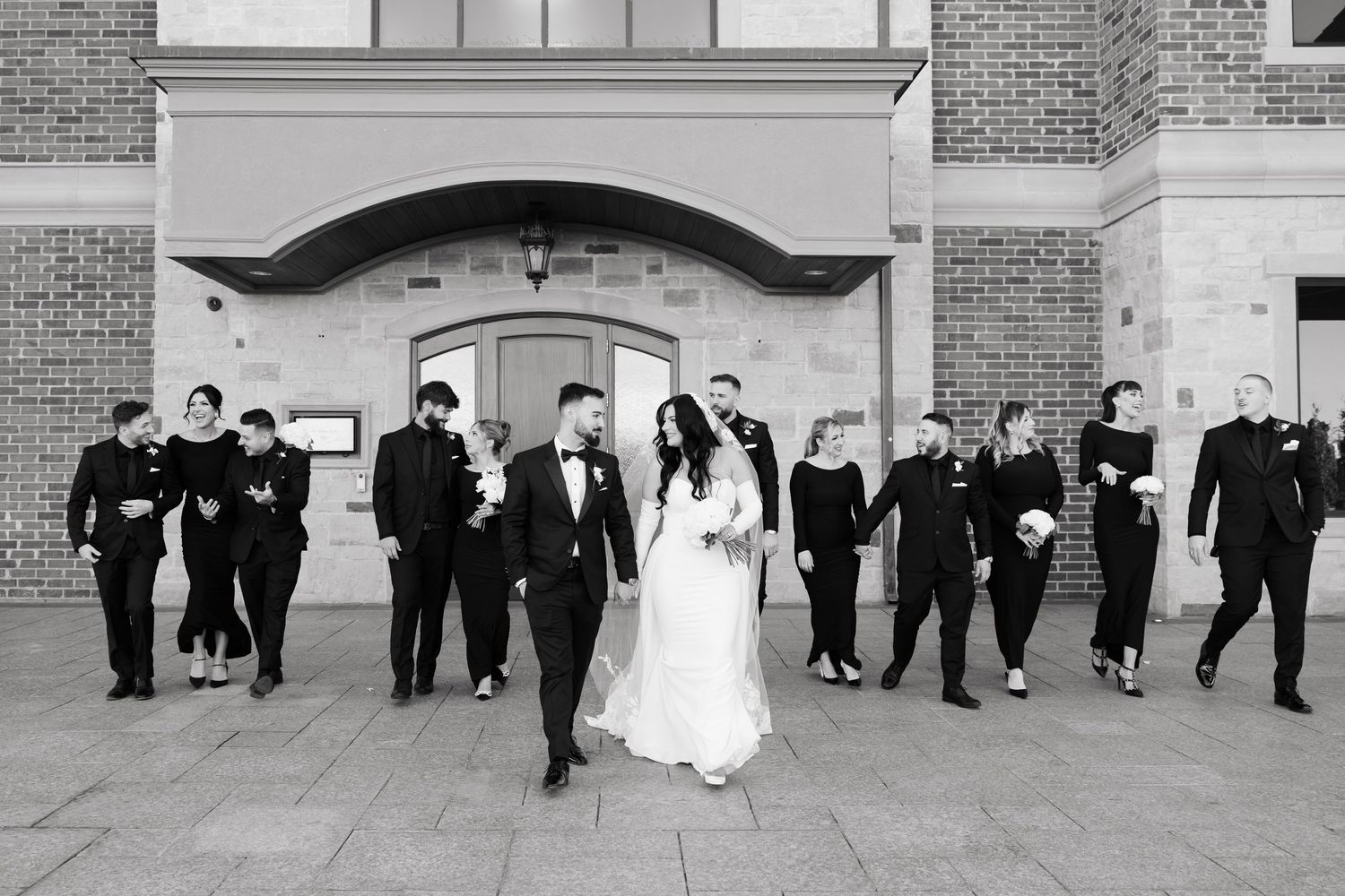 Wedding party in formal black attire walks alongside newlyweds outside elegant brick building.
