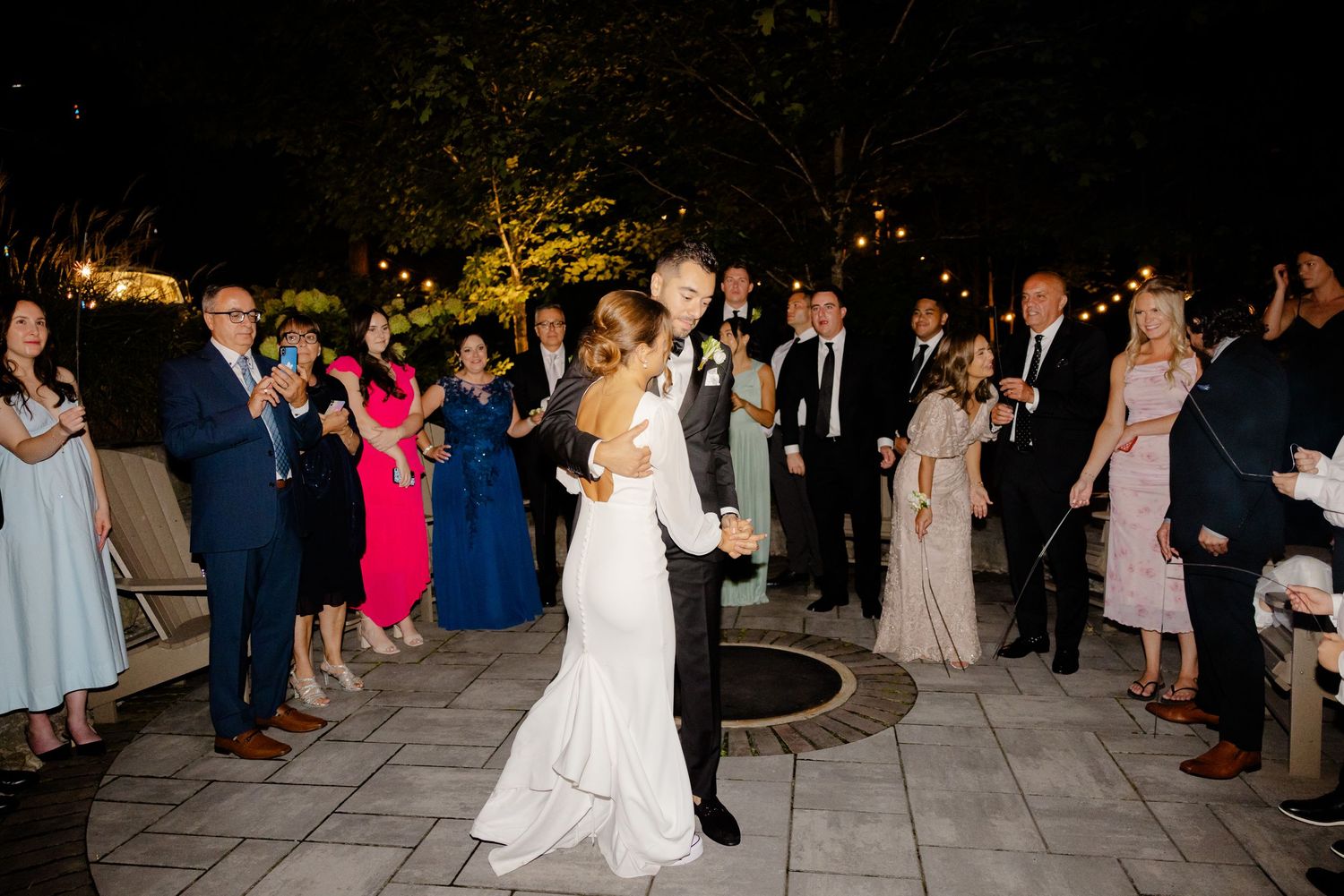 Wedding guests watch and cheer as the newlyweds dance on an illuminated stone patio.