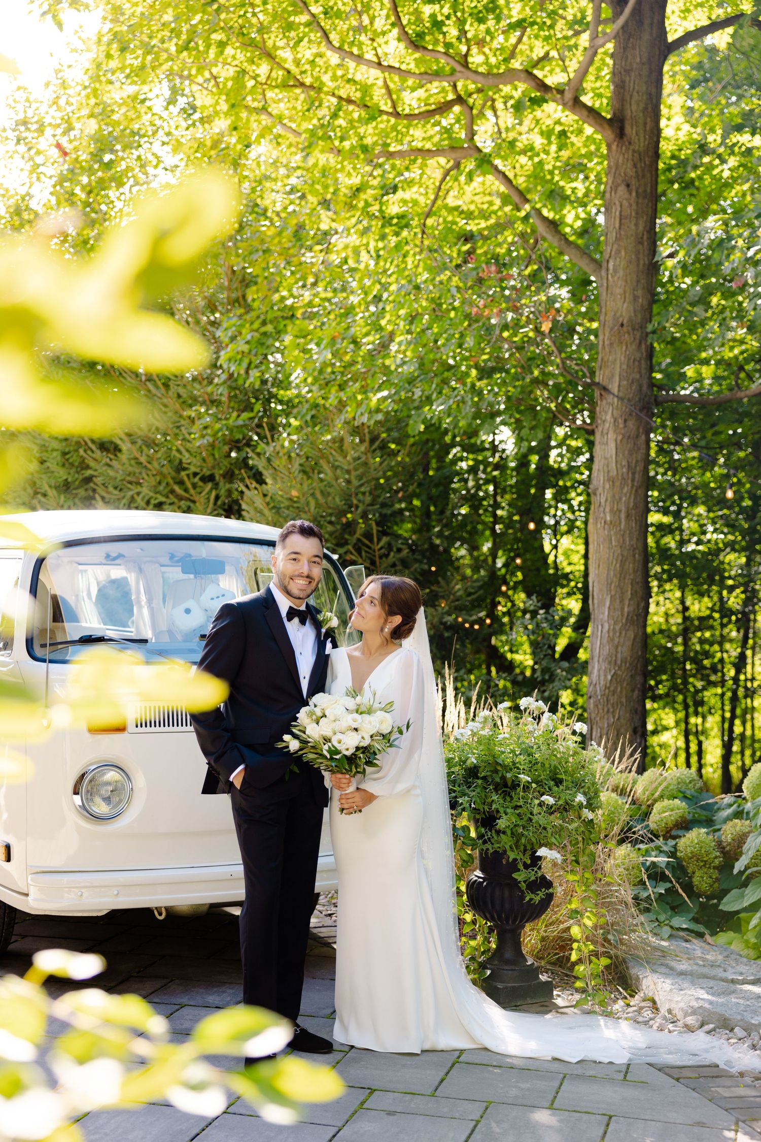 A wedding couple poses beside a vintage white van in a garden setting with soft sunlight filtering through trees.
