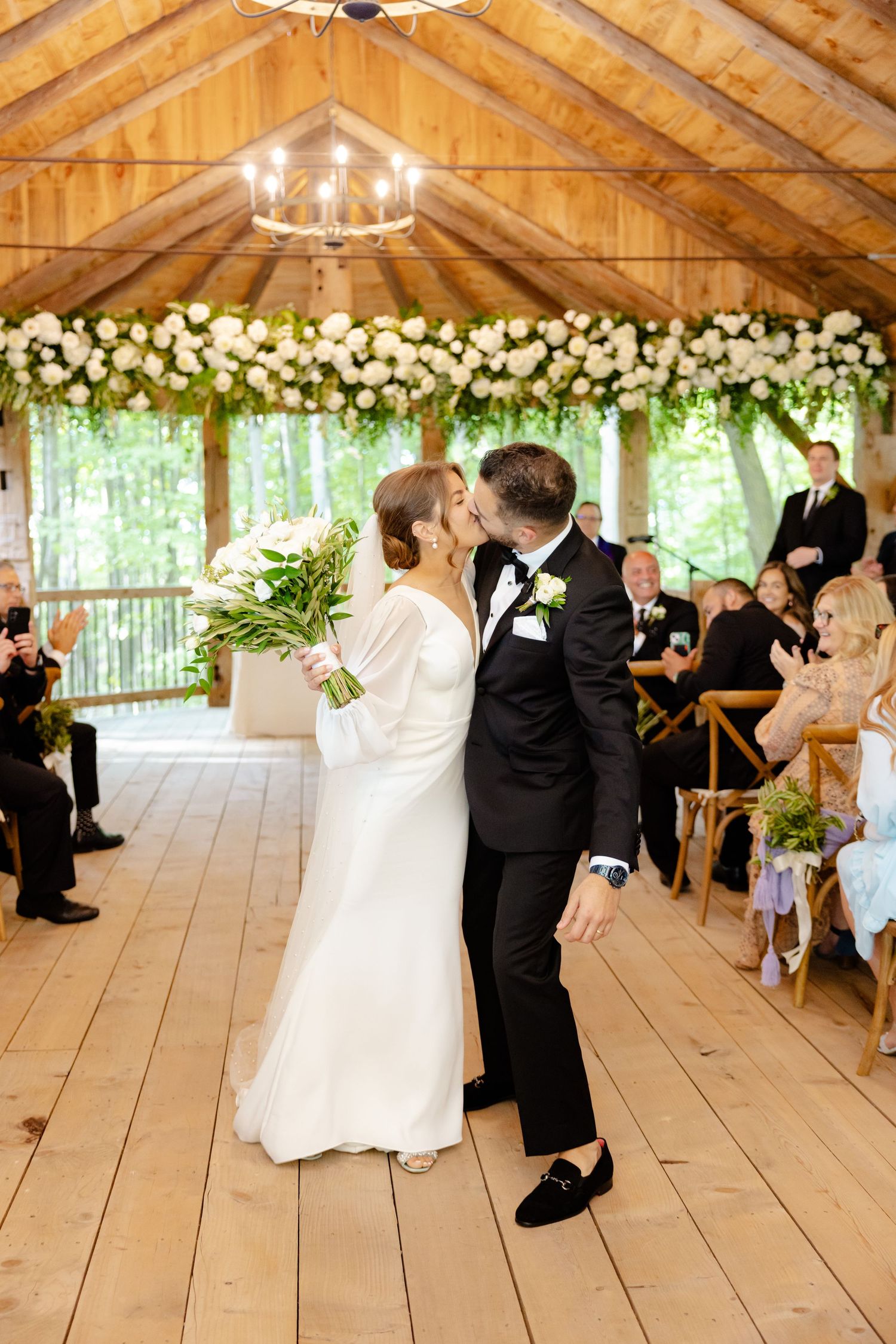 Intimate wedding moment in a rustic pavilion with white floral arrangements and natural backdrop.