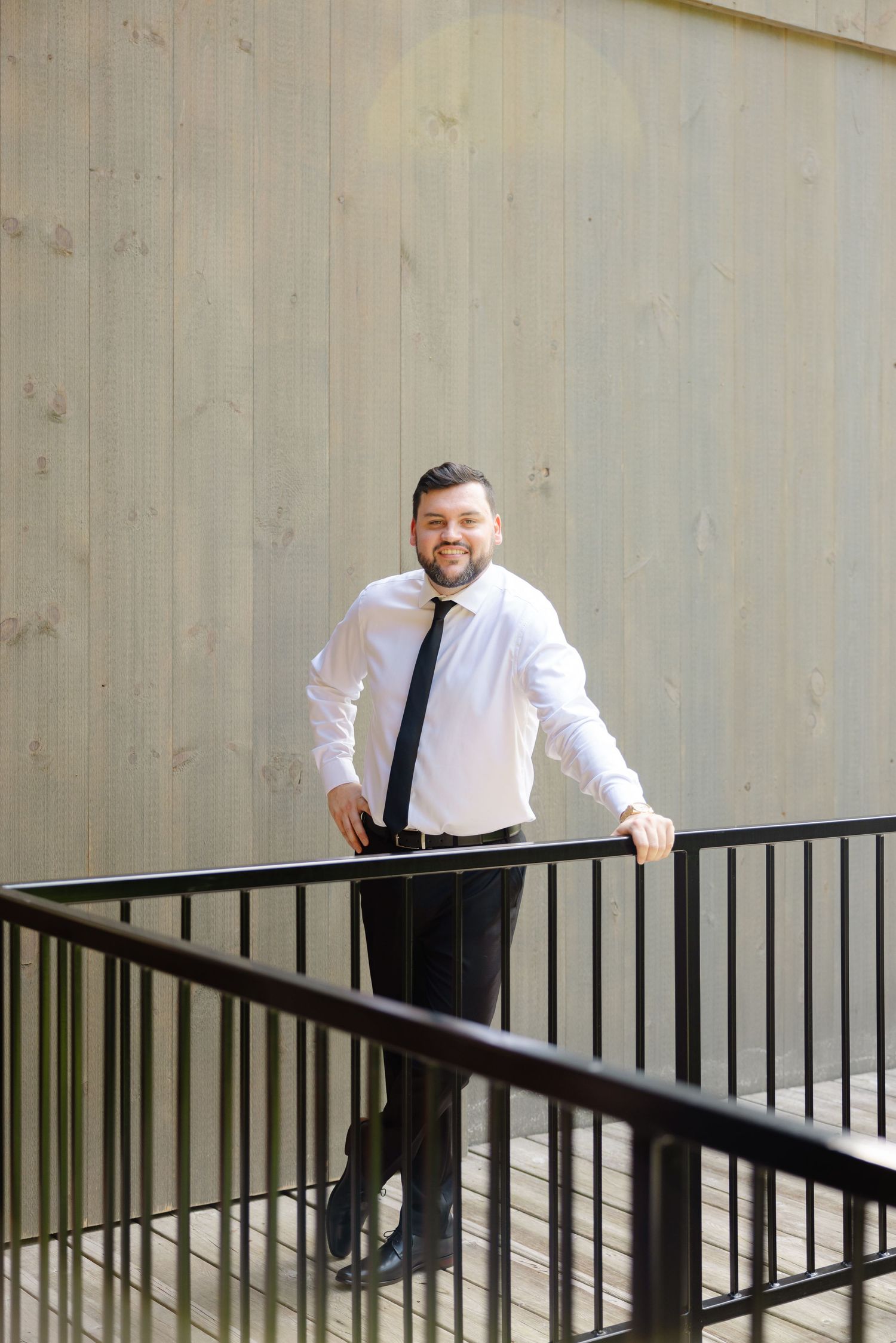 Person in white dress shirt and tie smiles while standing by railing on outdoor deck.