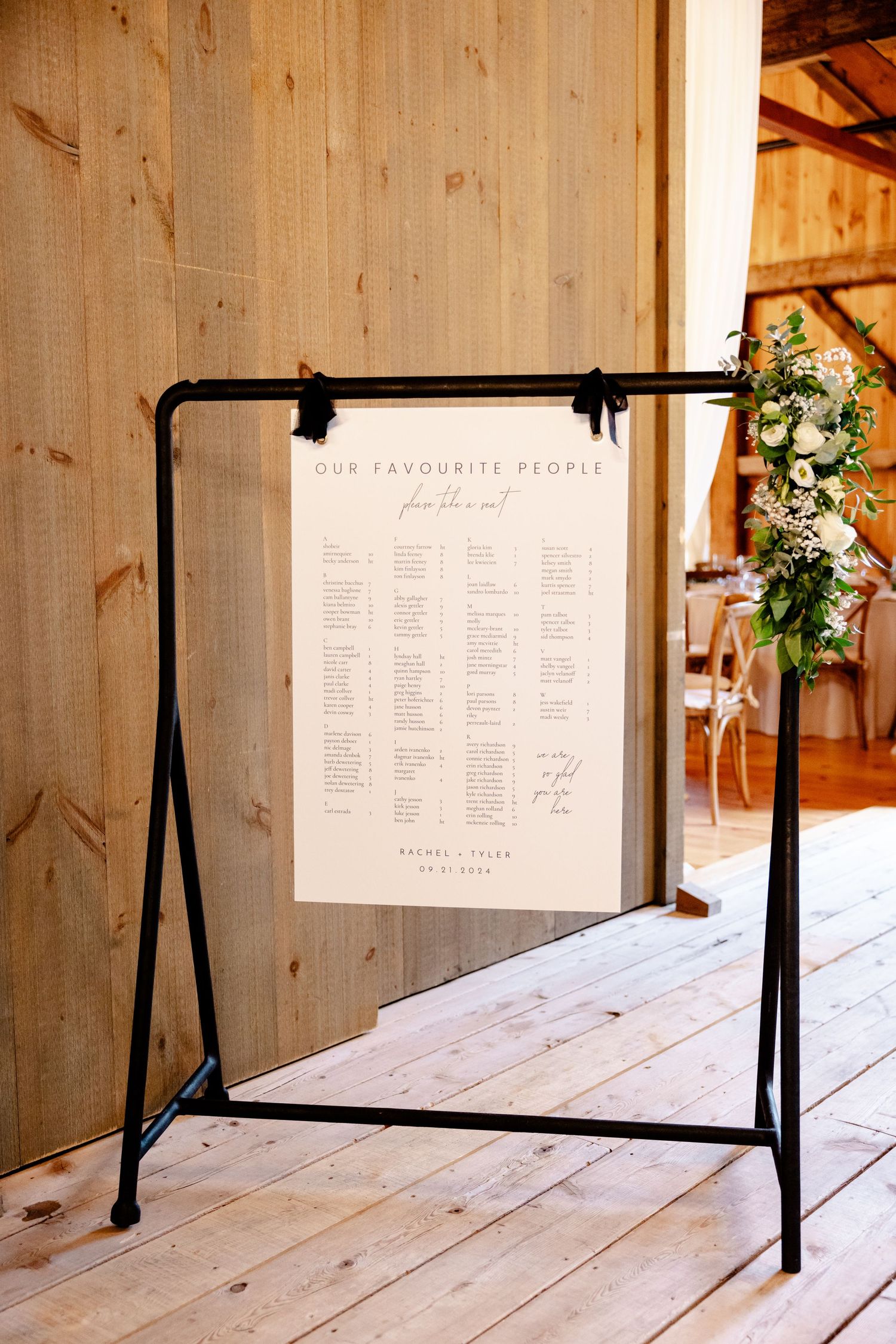 Wedding seating chart displayed on black metal stand frame against wooden barn wall.