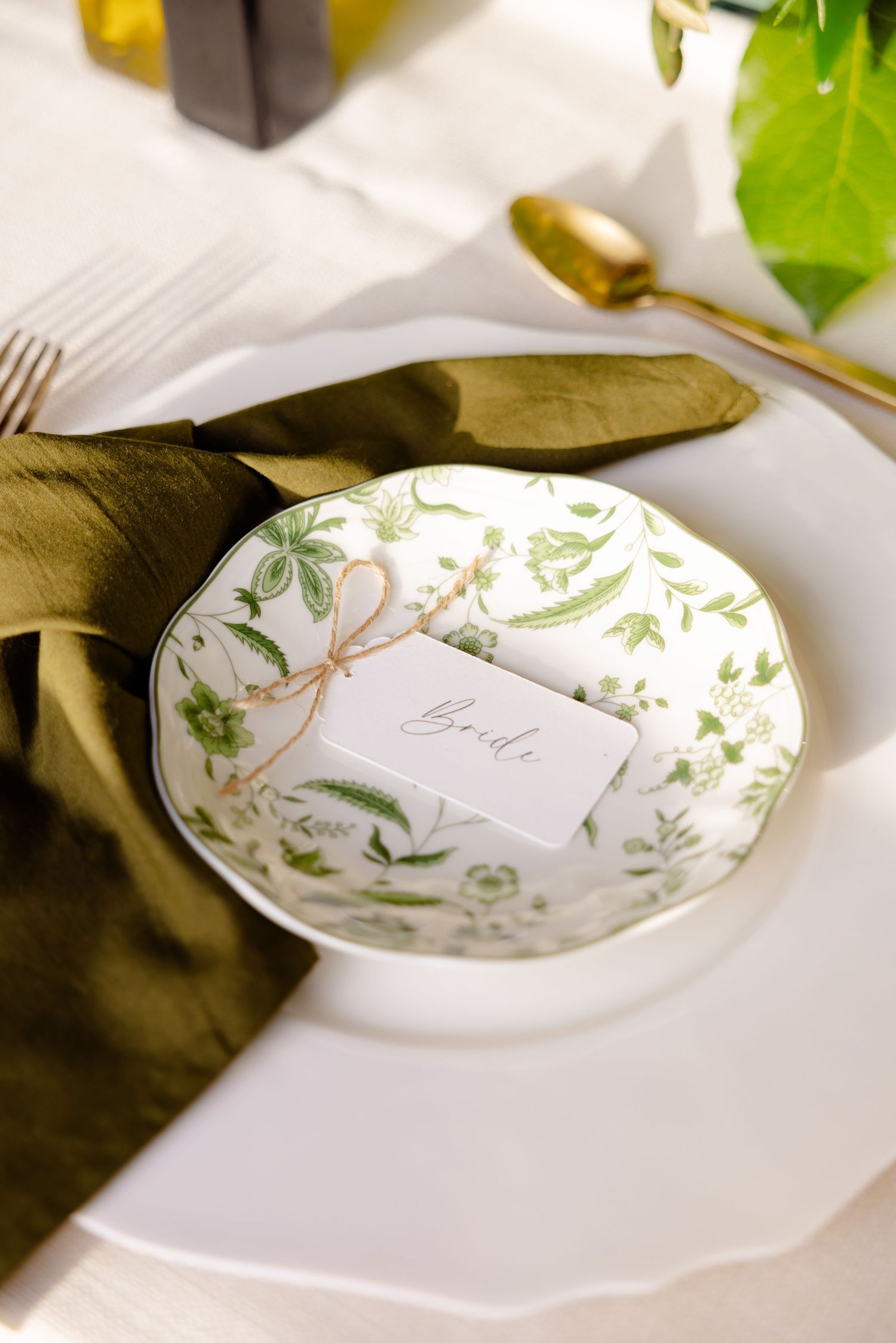 Close-up of place setting with green floral patterned plate and olive napkin on white tablecloth.