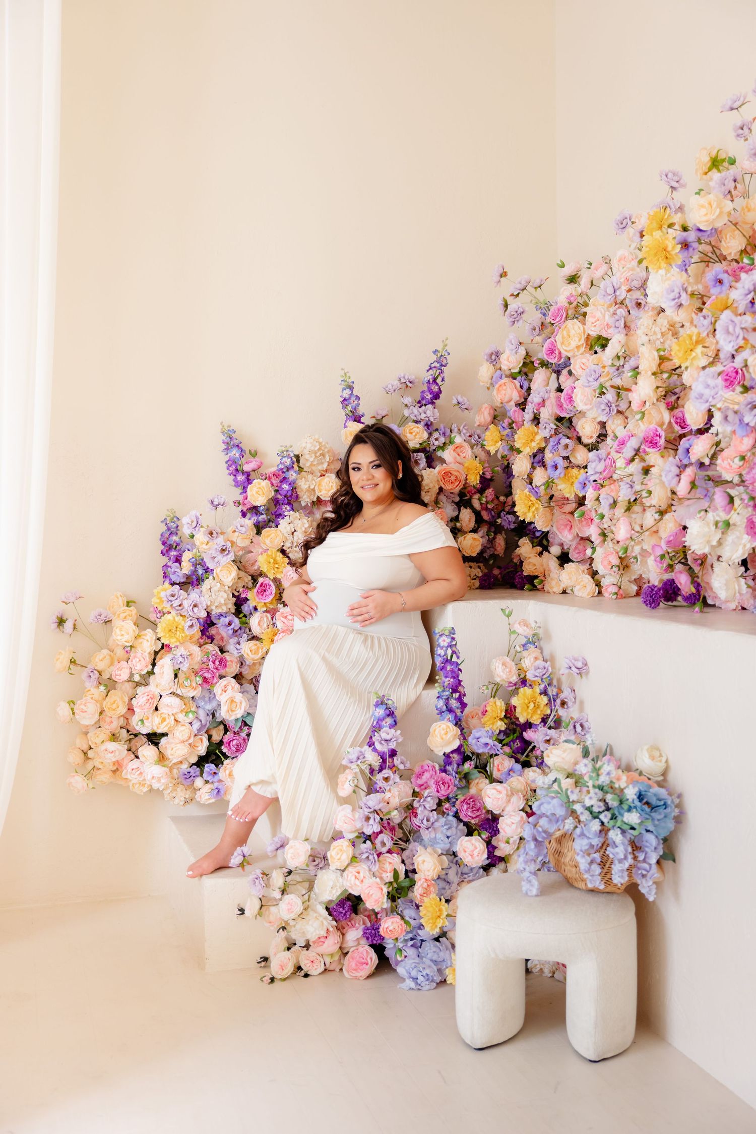 Maternity photo with flowing white gown against luxurious wall display of purple, pink and yellow flowers.