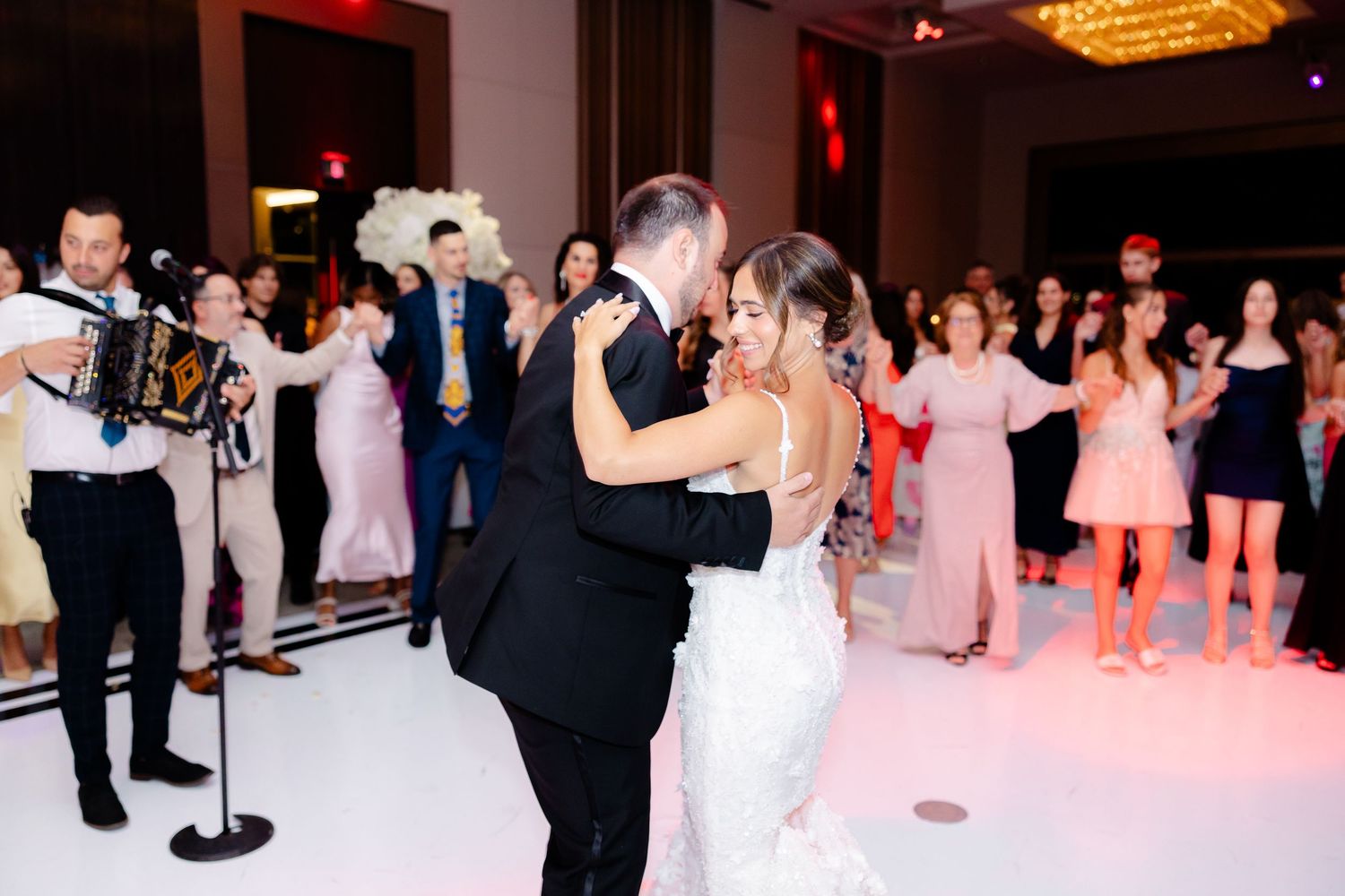 Multiple couples dancing together at an elegant wedding reception with red lighting and a crystal chandelier overhead.