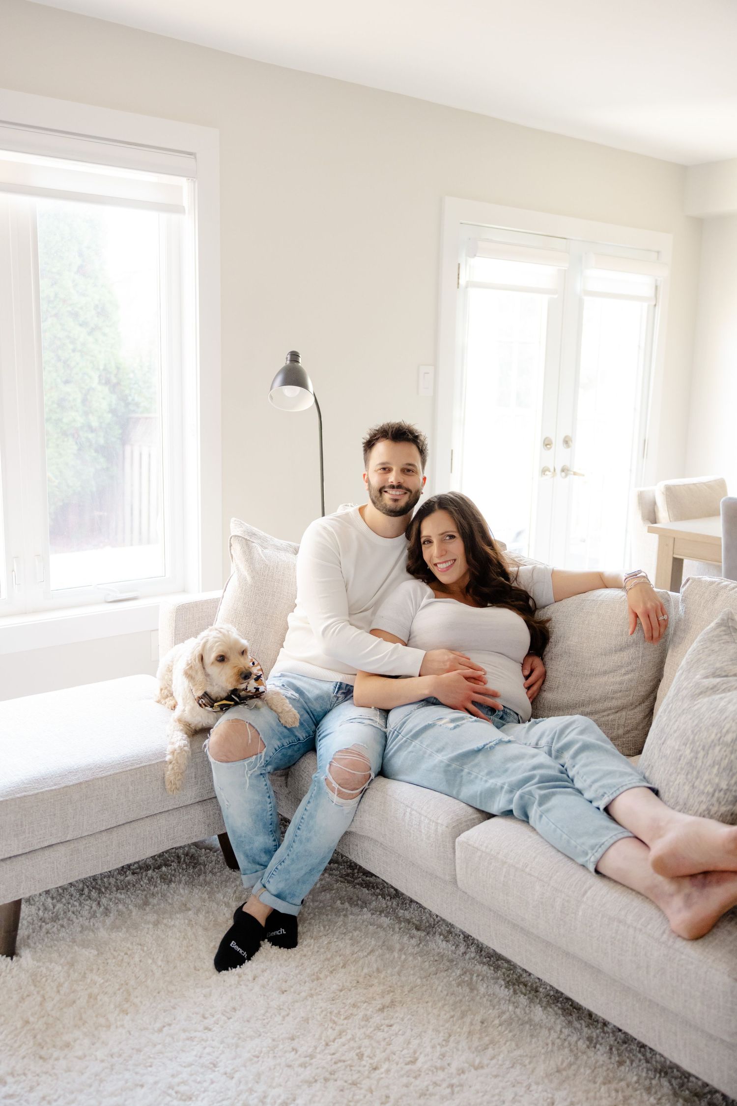 A couple and their dog relax on a couch in a bright living room with large windows.