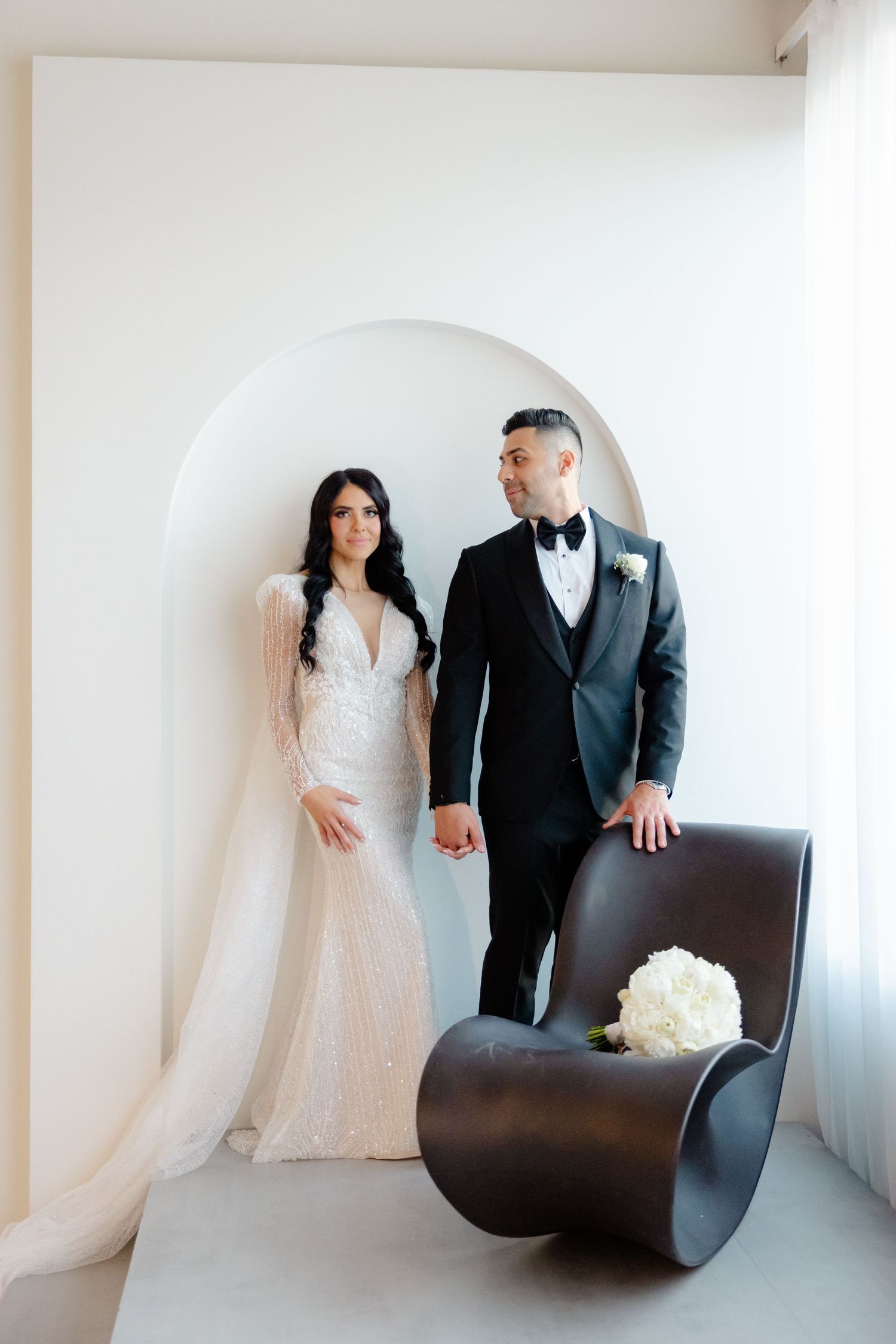 Modern wedding couple posing in white archway with contemporary curved chair and white bouquet.