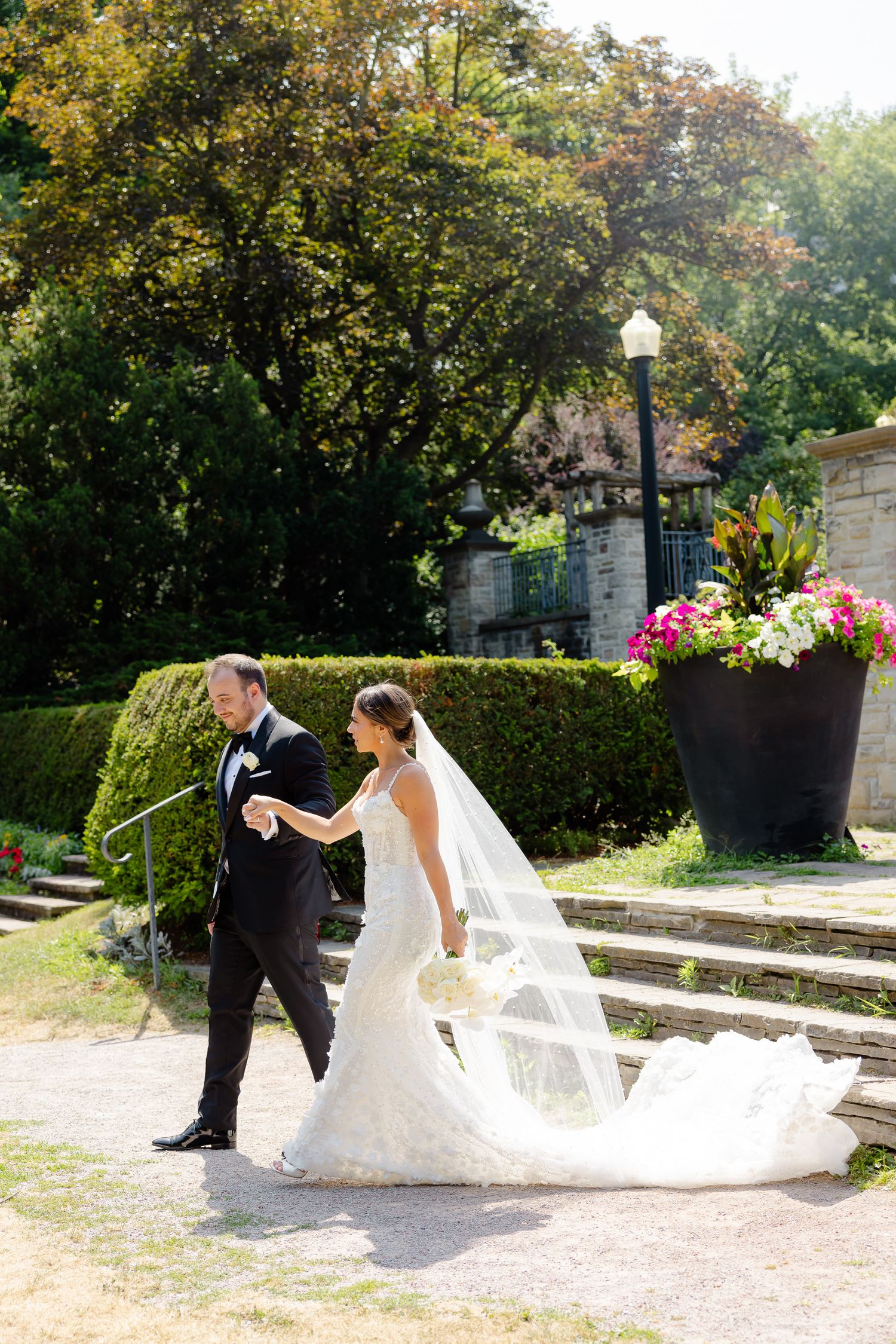 A couple in wedding attire walk through a garden path bordered by manicured hedges and flowering plants on a sunny day.