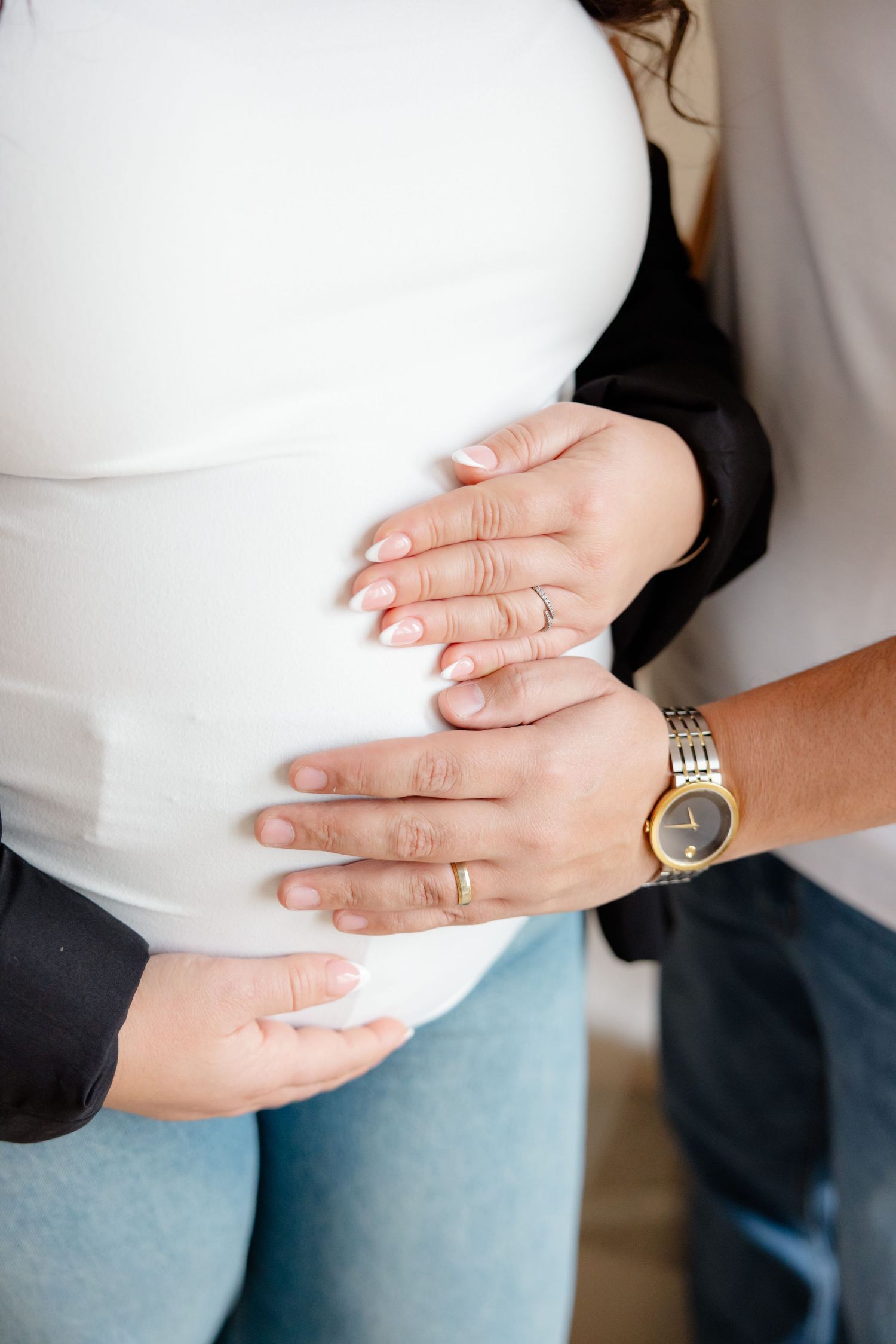 Hands gently rest on a pregnant belly, with one person wearing a gold watch and white shirt against light denim jeans.