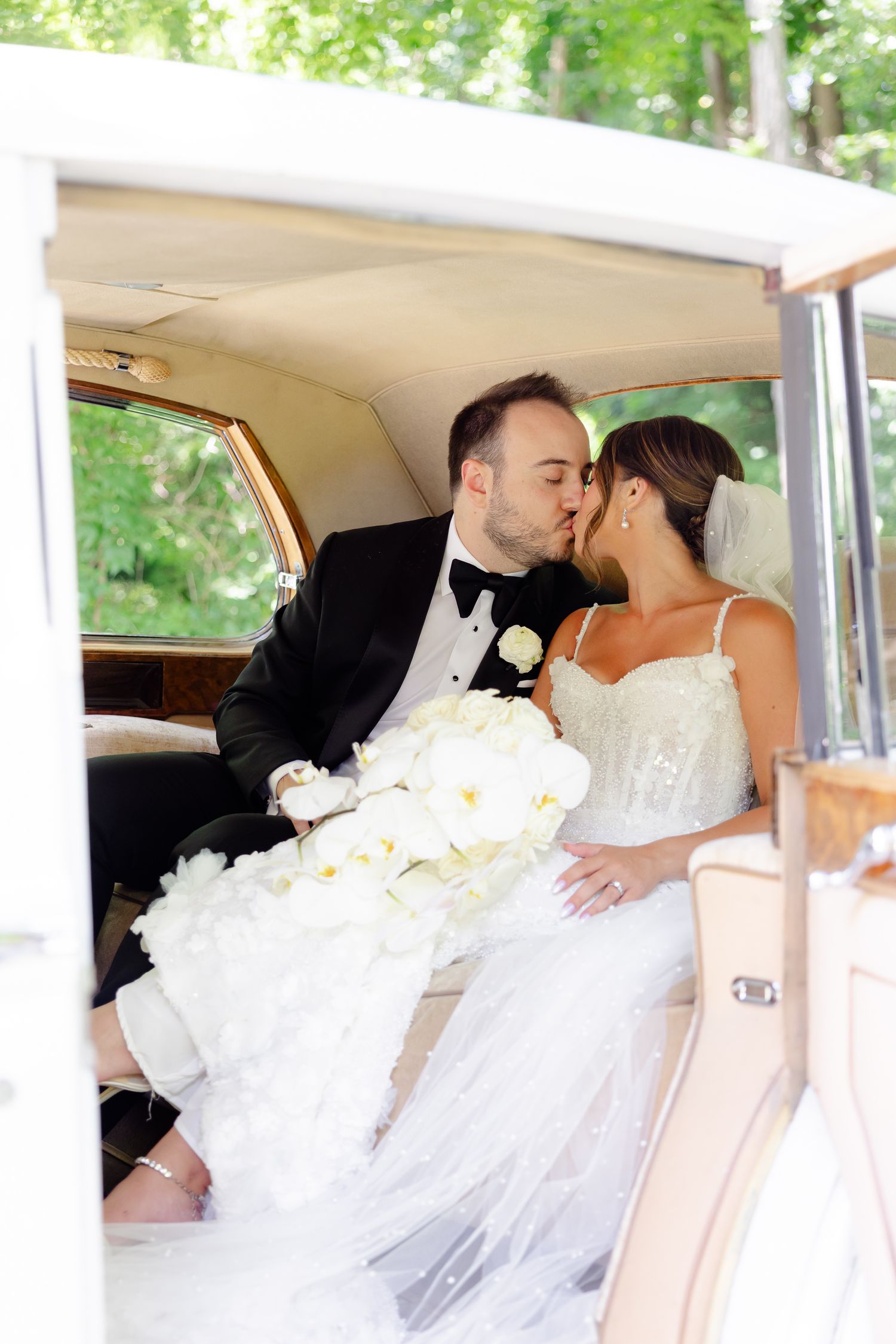 A romantic wedding couple shares tender kisses while seated in the back of a vintage car.