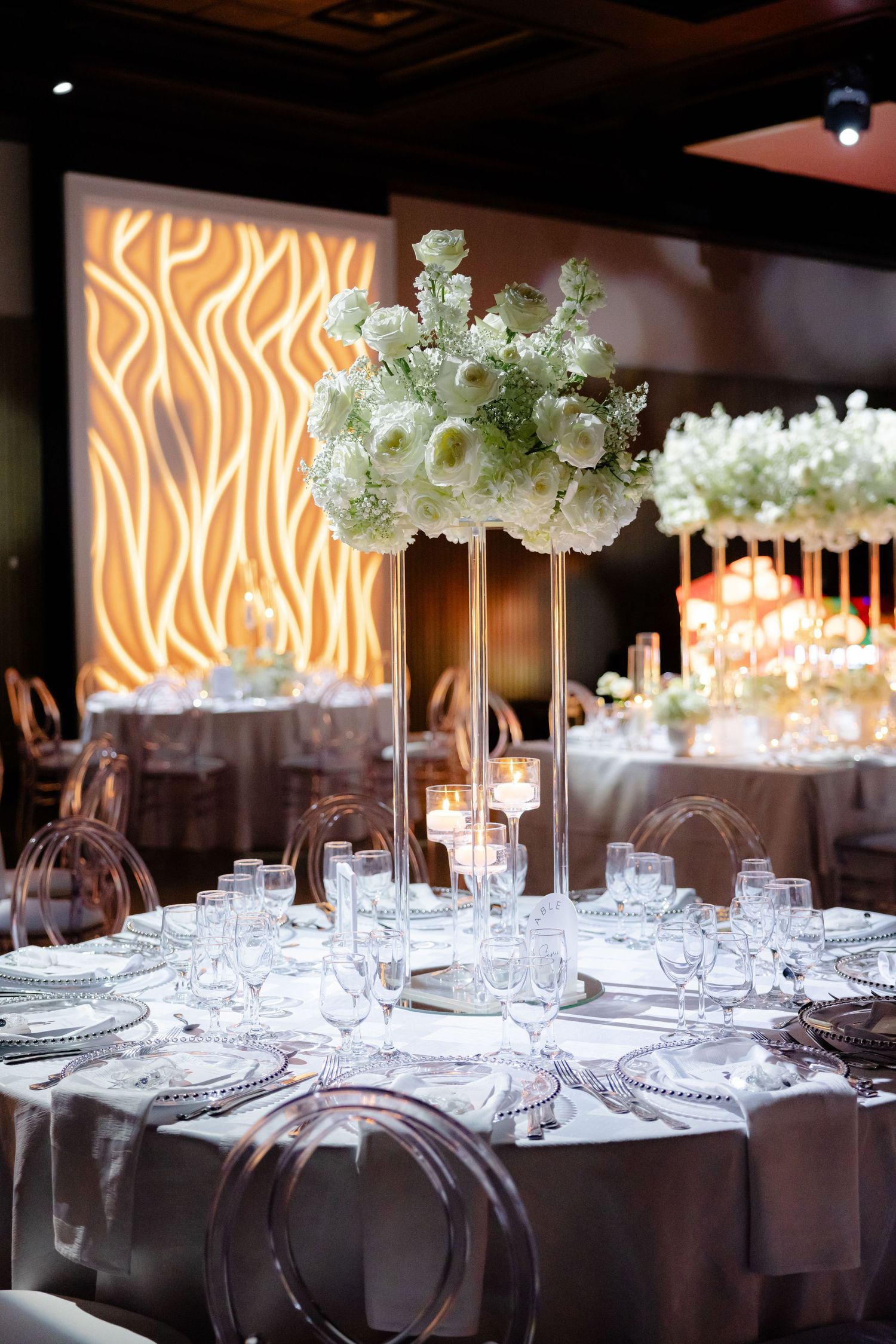 Modern wedding reception table with tall white floral centerpiece against illuminated geometric wall design.