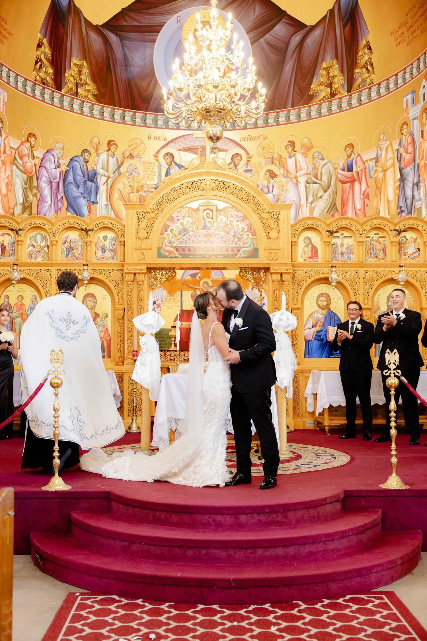 Orthodox wedding ceremony taking place on red carpet steps inside ornate church with golden icons and chandeliers.