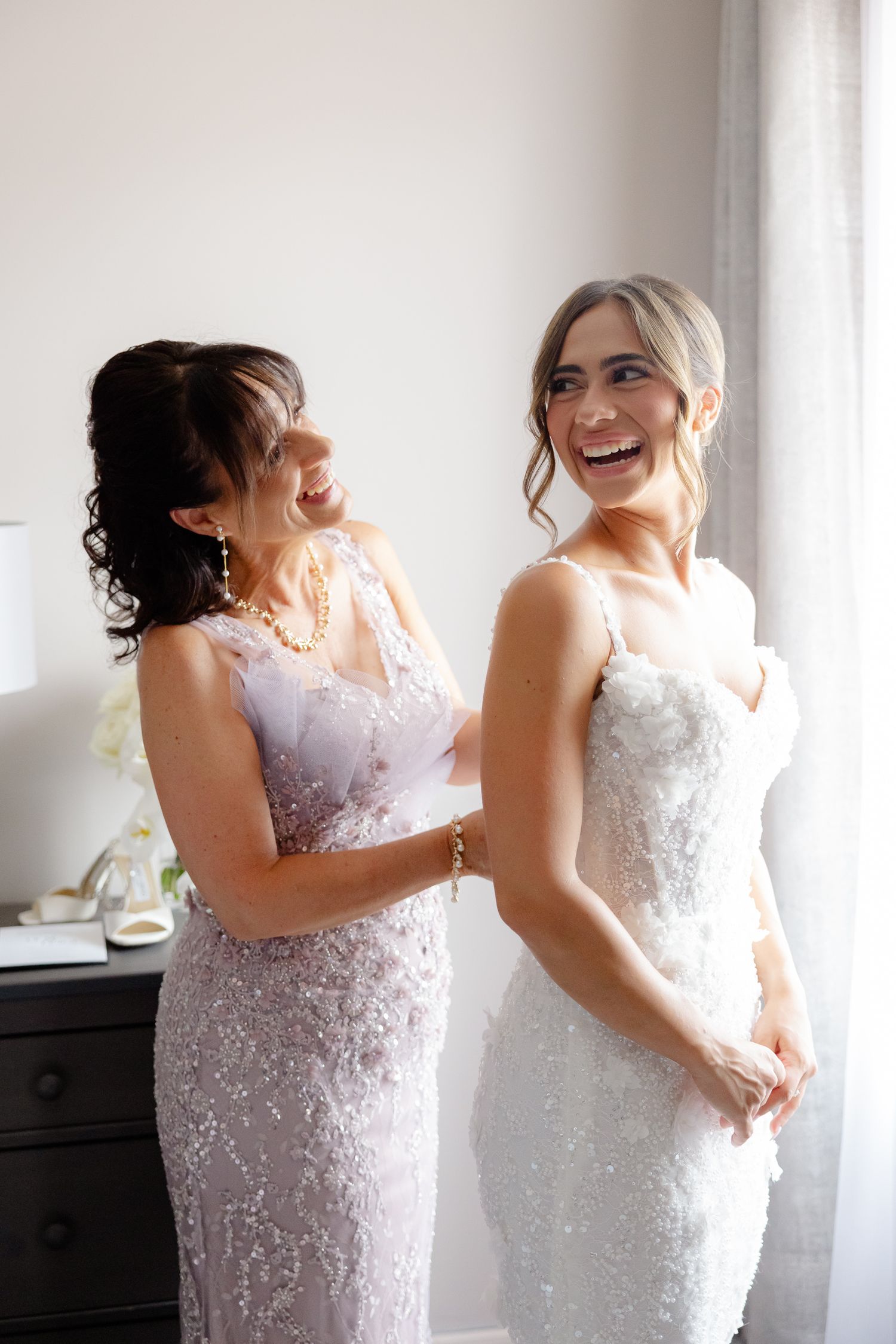 Two women share a joyful moment wearing elegant lace dresses in white and blush tones by a window.