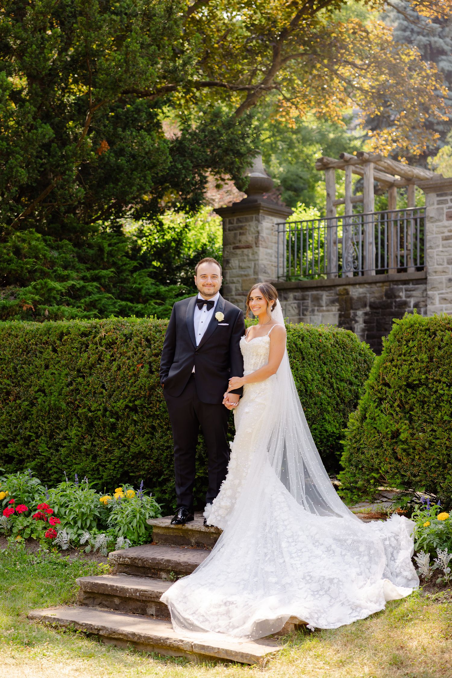 A bride and groom pose together on stone steps surrounded by blooming gardens and elegant landscaping.