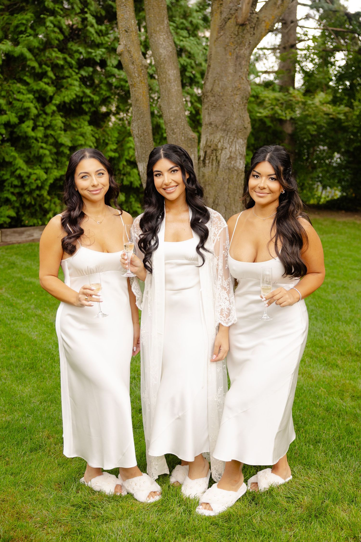 Three women in white dresses pose playfully together on grass near trees in an outdoor setting.