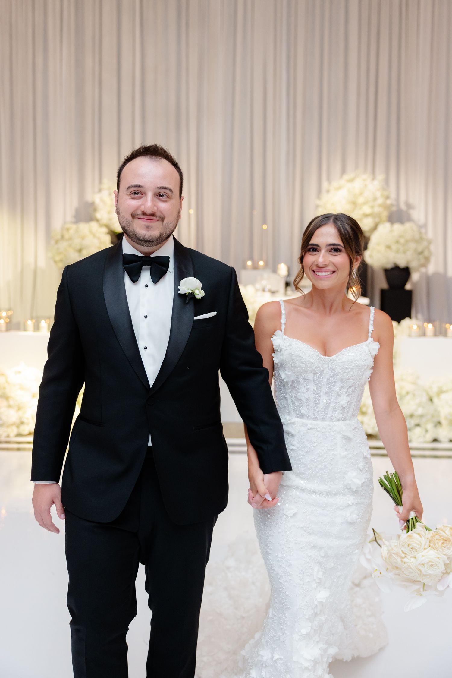 A bride and groom hold hands and smile while walking at their elegant wedding reception with white floral decor.