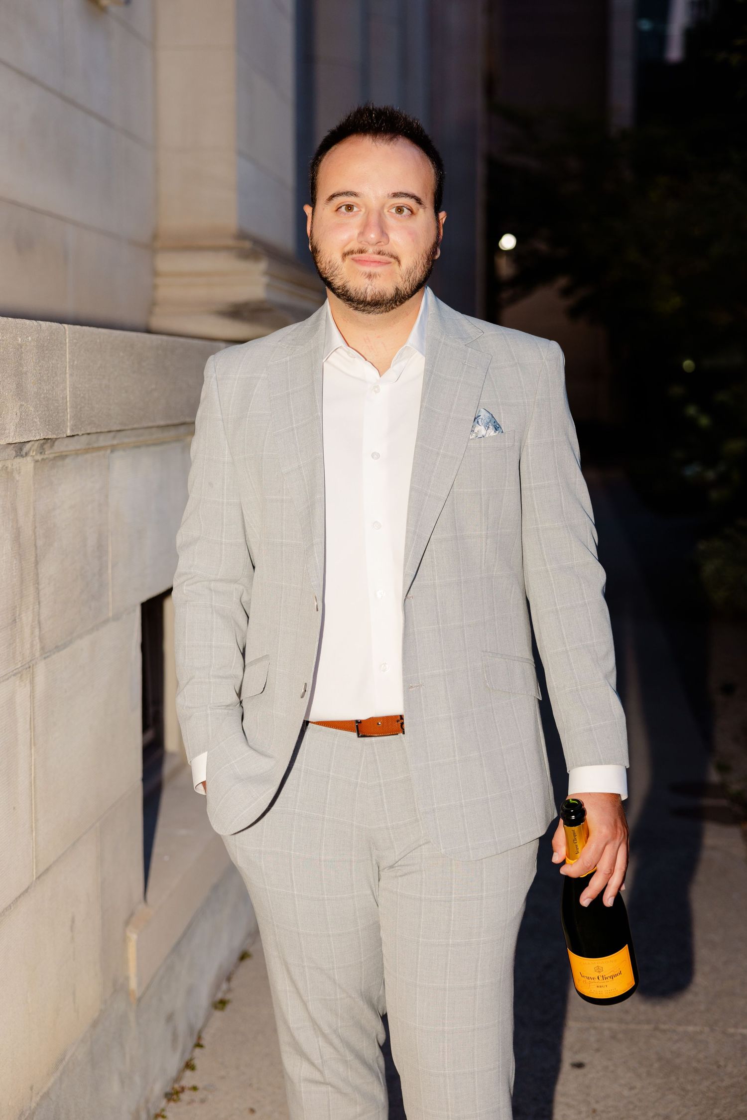 Man in light gray suit and white shirt holding champagne bottle against stone building.